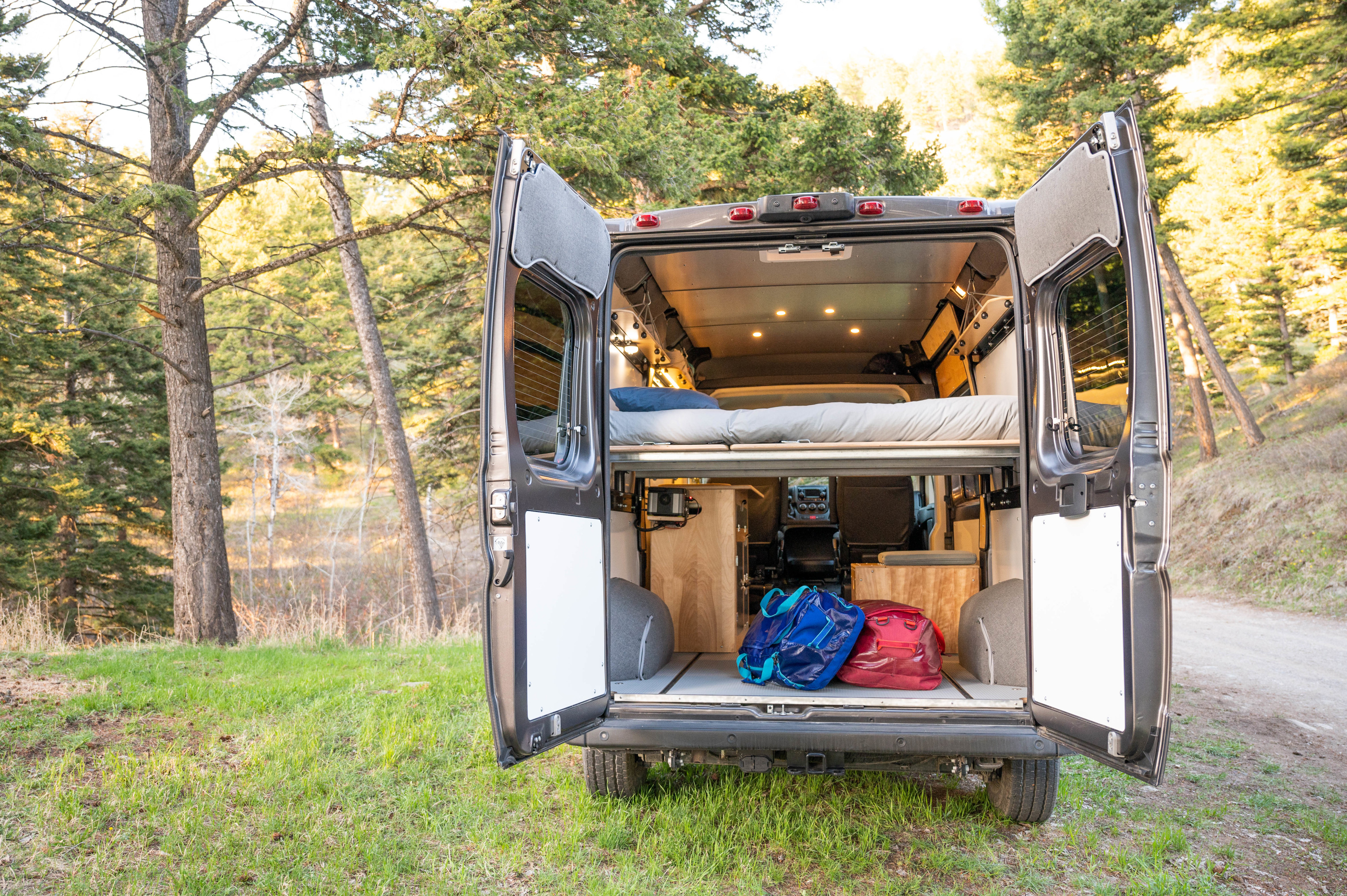 Open rv rental camper van rear view. Shows cozy bed, storage with bags, and illuminated interior, parked in a forest.. RAM Promaster 1500 2021