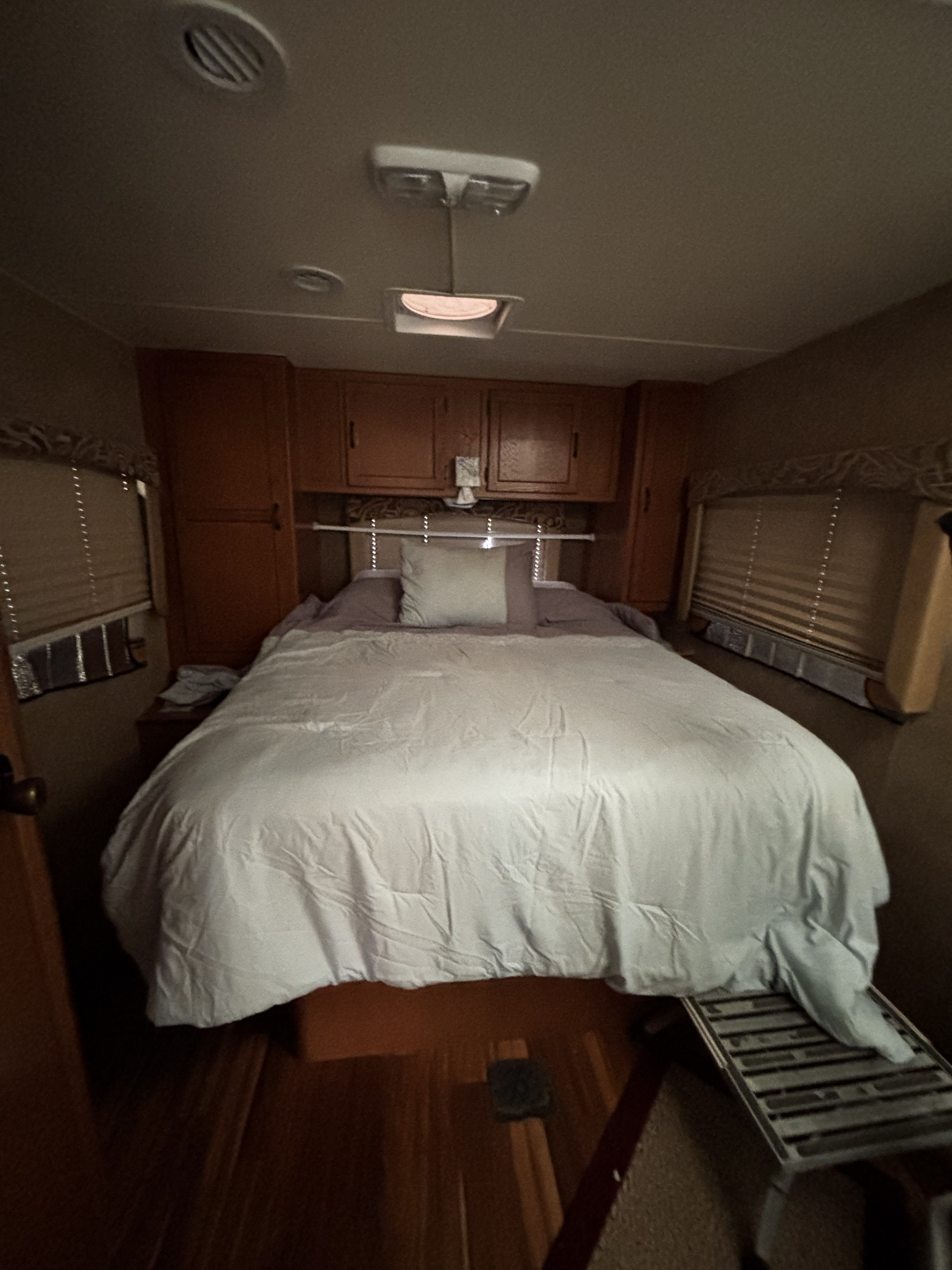 Cozy RV rental bedroom featuring a large bed, wooden cabinets, windows with blinds, ceiling vents, and an access step.. Chevy Express 2013