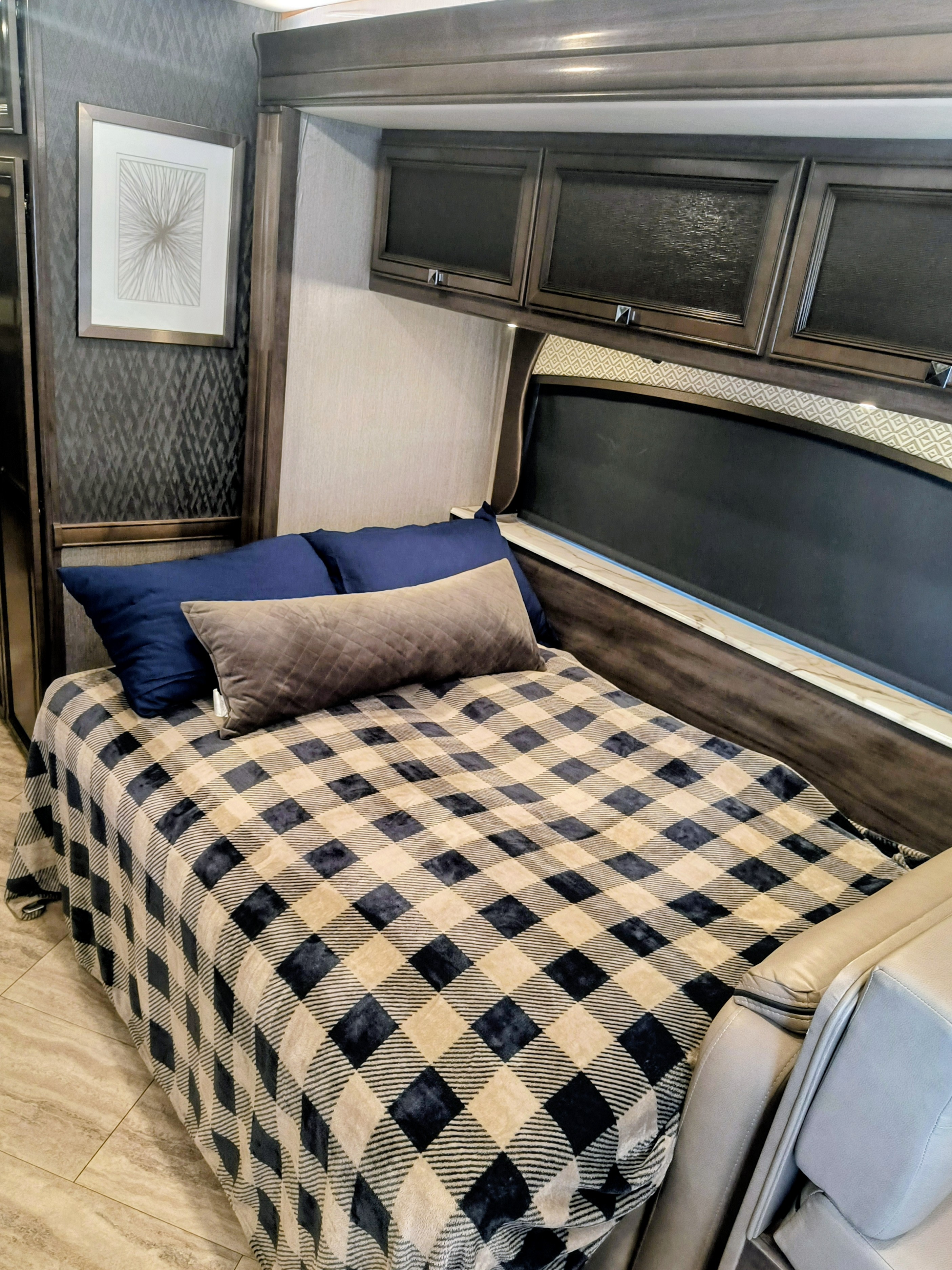 Cozy RV rental bedroom featuring a bed with a dark plaid blanket, blue & gray pillows, dark wood cabinets, and framed wall art.. Fleetwood Fortis 2020