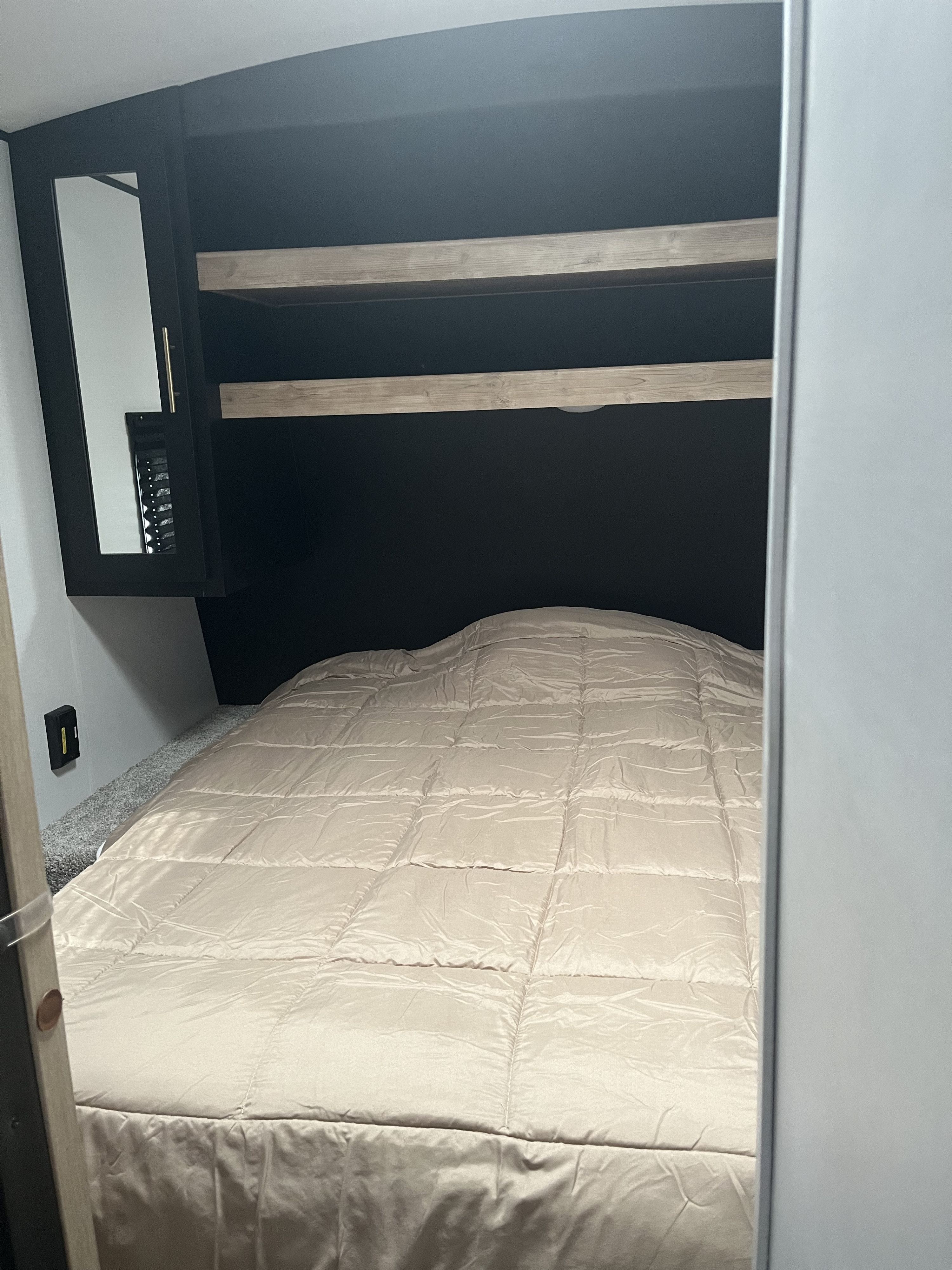 RV rental bedroom interior: a made bed with a light comforter, overhead shelves, and a small mirrored cabinet.. Keystone RV Springdale 2022