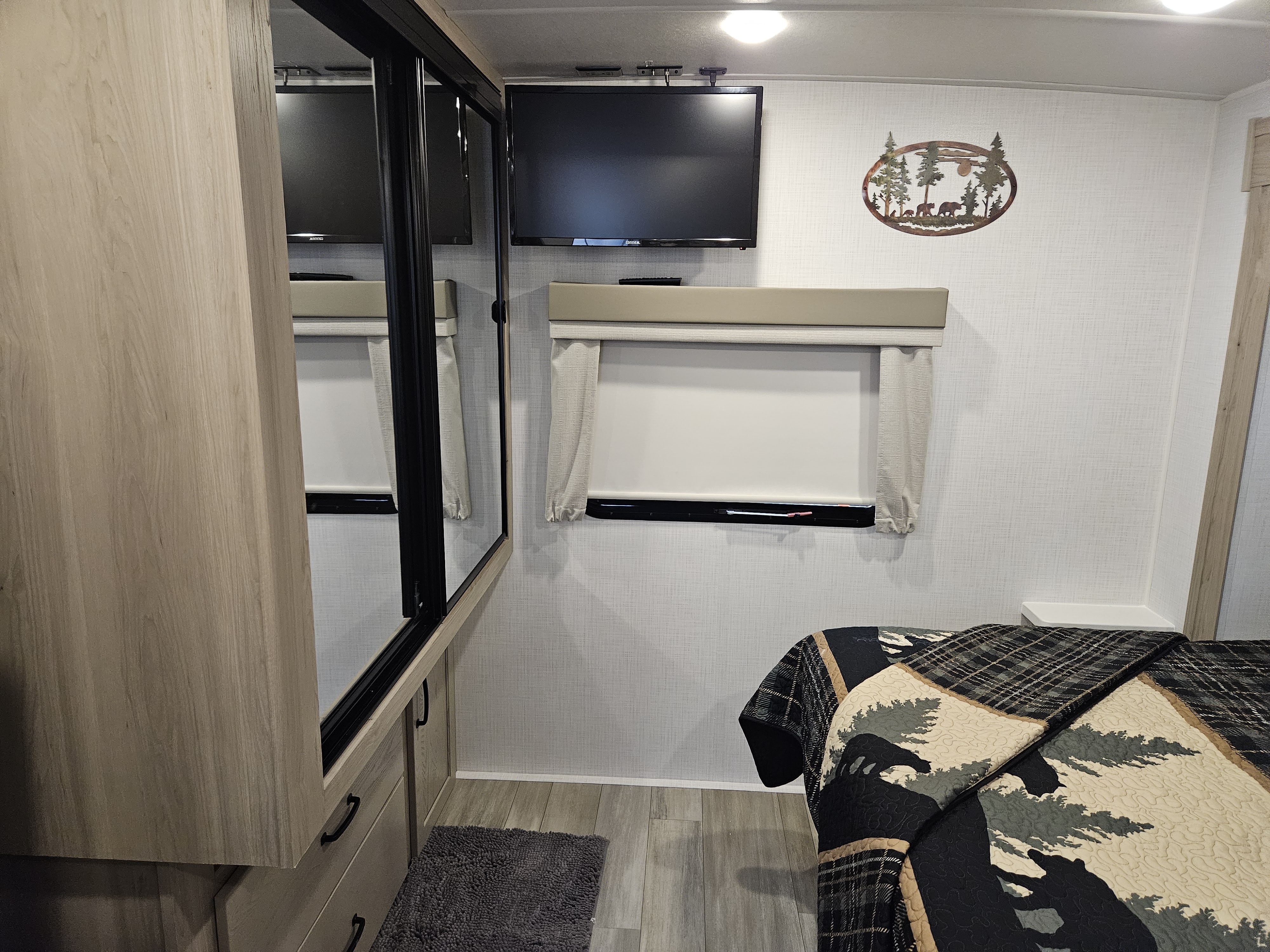 Inside an RV rental bedroom: a TV, window, forest art, and bear-themed bedspread. Mirrored wardrobe doors and a small rug add to the space.. Forest River Flagstaff Micro Lite 2024