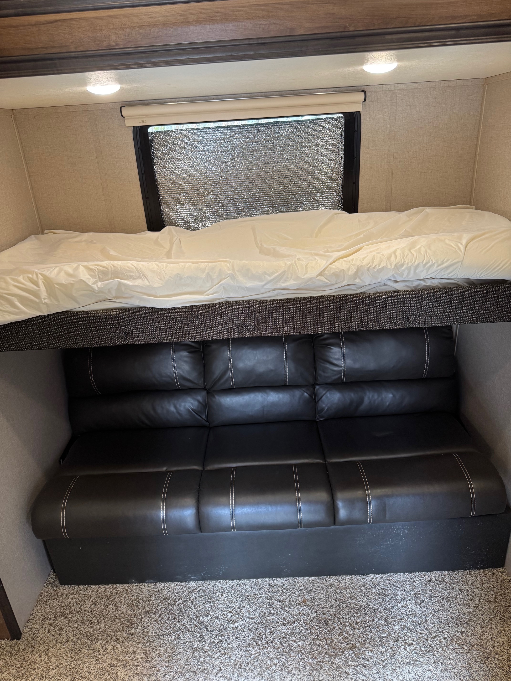 RV rental interior: bunk bed with white bedding above a black faux leather sofa. Window covered with reflective material for privacy.. Forest River Coachmen Chaparral 2020