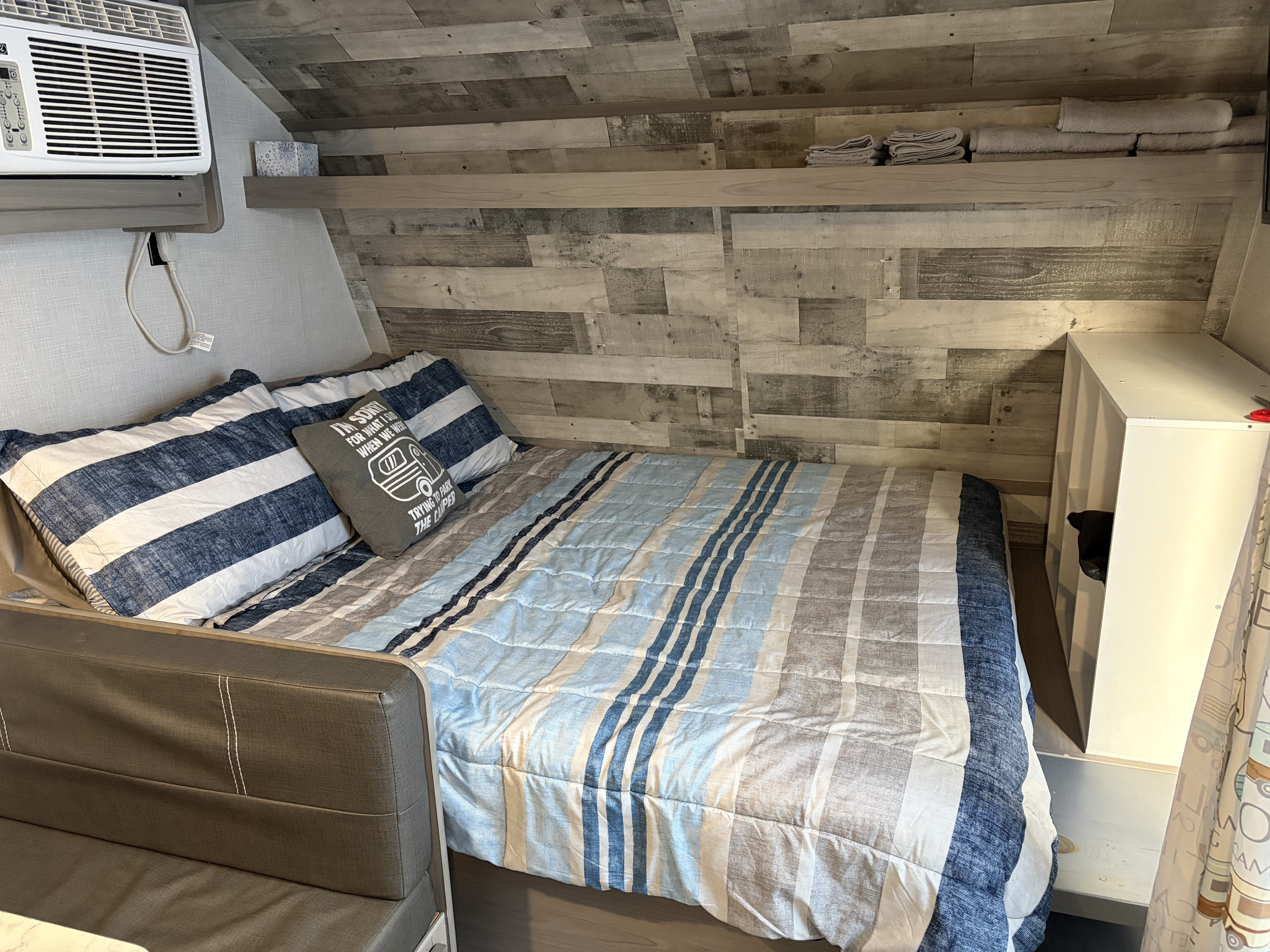 An inviting RV rental interior shows a bed with blue/white striped bedding, a humorous camper pillow, and an AC unit.. Dutchmen Coleman Lantern LT 2022