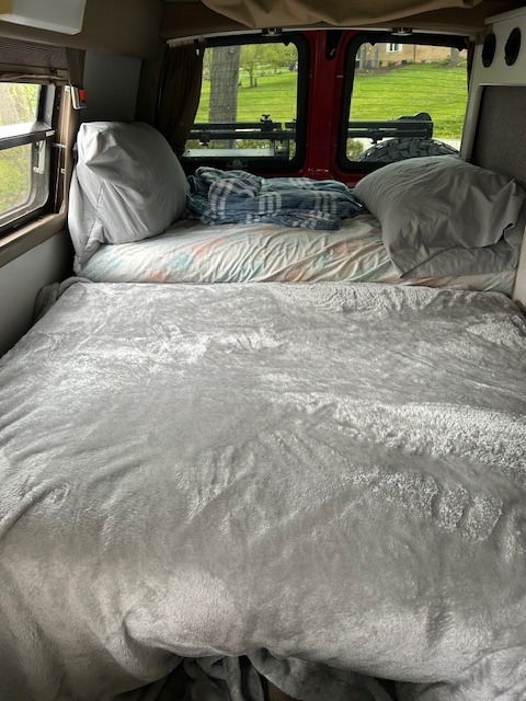 Cozy RV rental bed with a soft, grey blanket, pillows, and an inviting green outdoor view. Ideal for your travel adventures.. Chevrolet Express Silverado 3500 4X4 Sportsmobile edition 2008