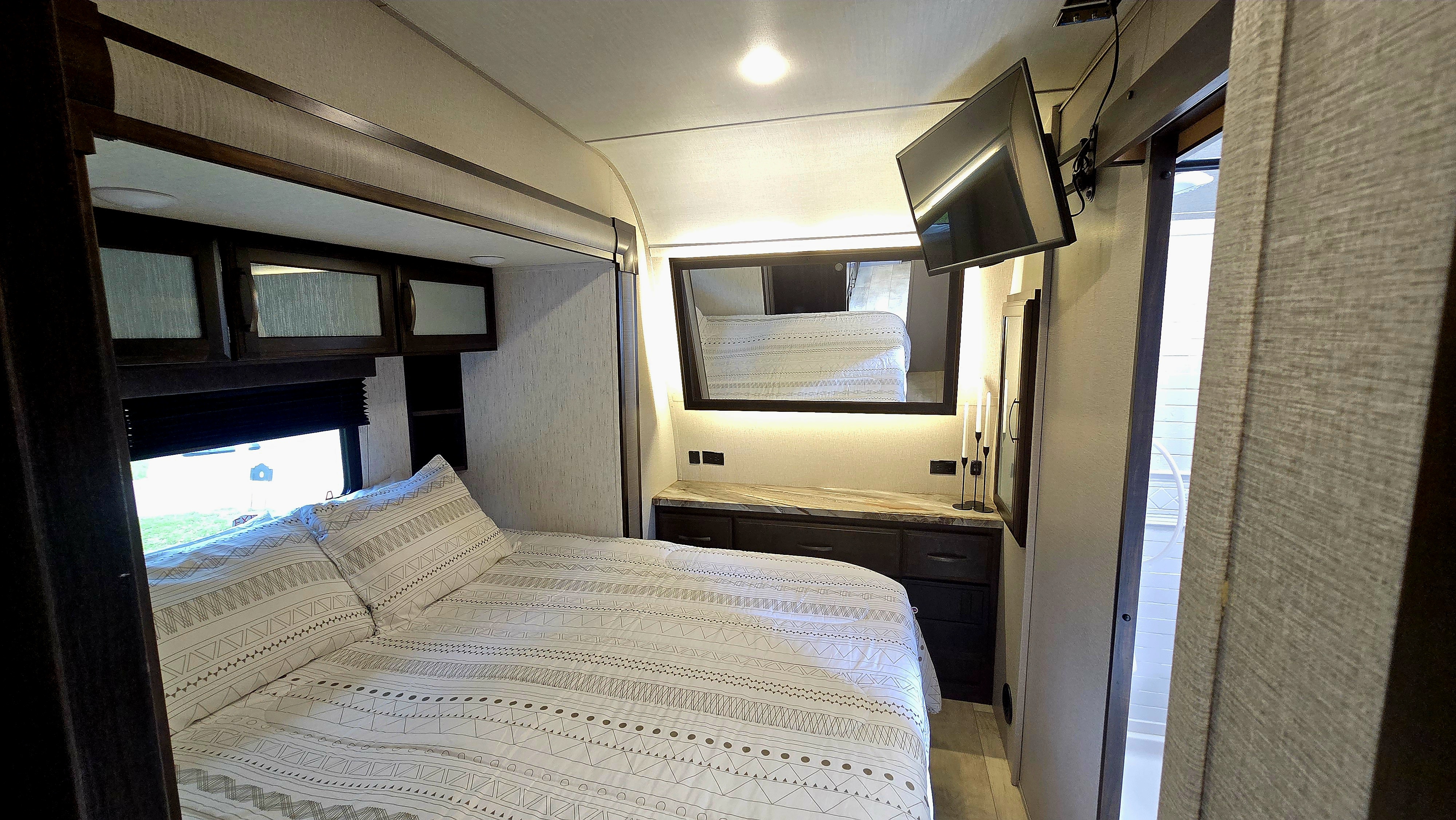 RV rental bedroom with patterned bed, window view, overhead storage, dresser with mirror & TV. Comfortable travel lodging.. Grand Design Momentum 2020