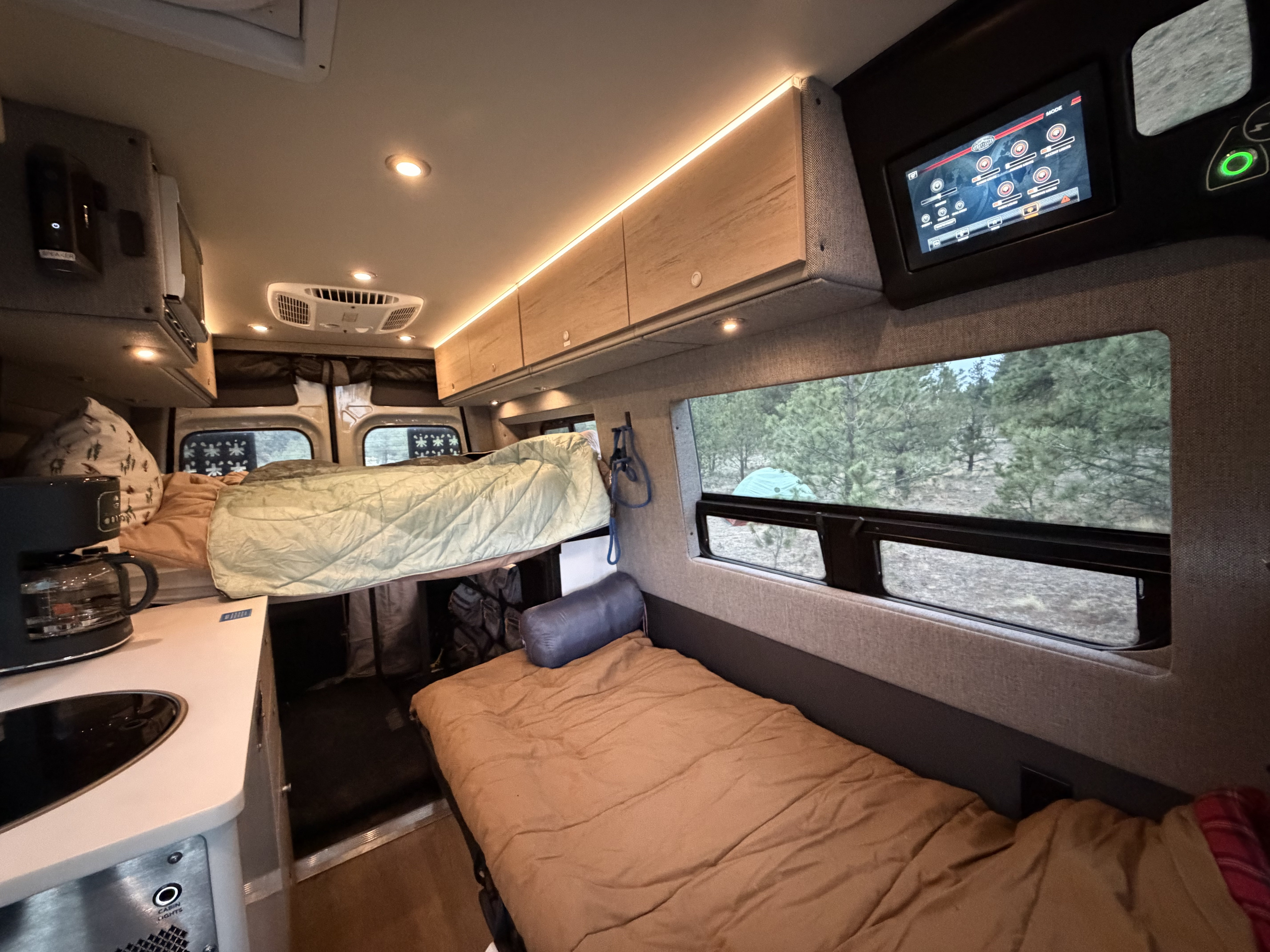 Cozy RV rental interior: an elevated bed, lower sleeping area, kitchenette, and control screen, all with a serene forest view.. Mercedes MB 212 Sprinter 2025