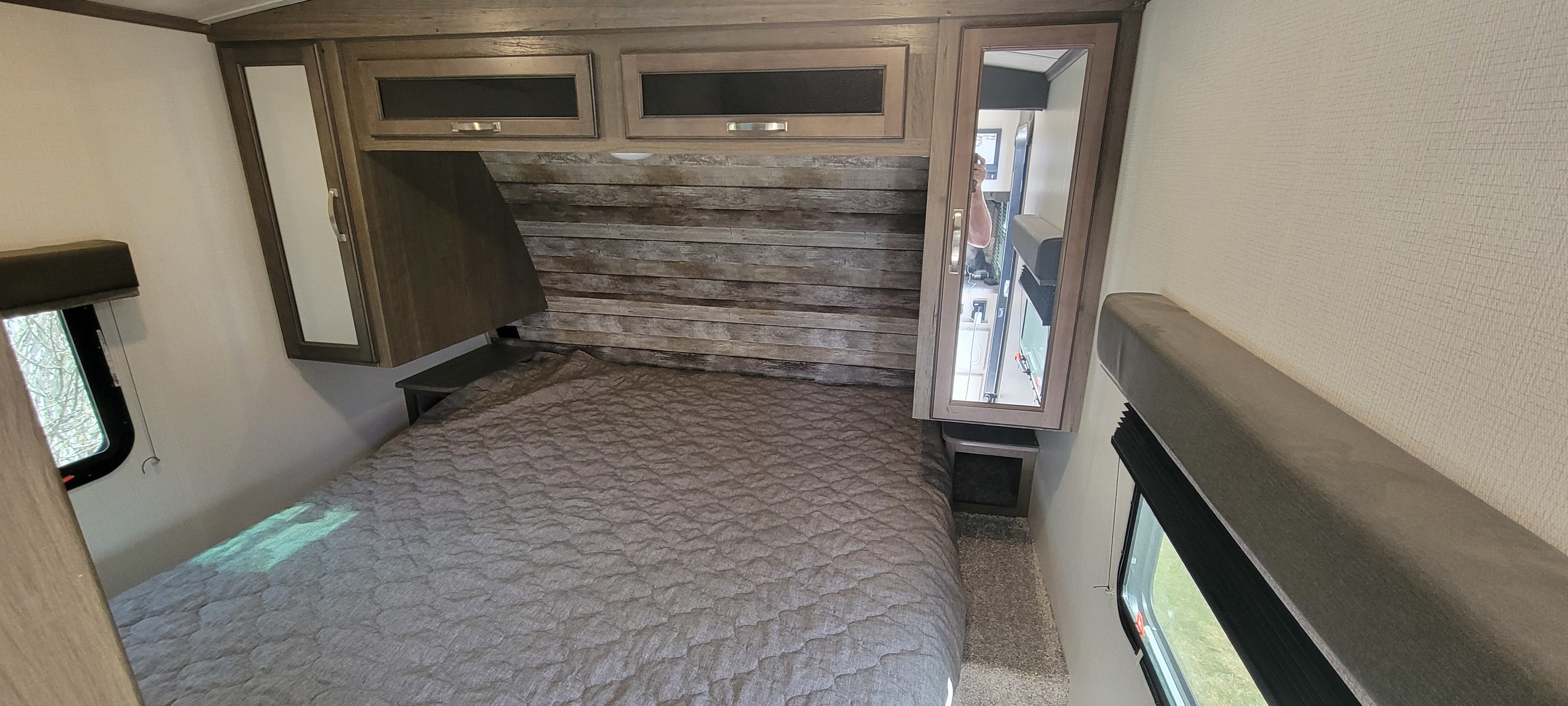 This RV rental bedroom features a gray bed, rustic wood headboard, overhead cabinets, windows, and a mirrored wardrobe.. Keystone RV Cougar 2021