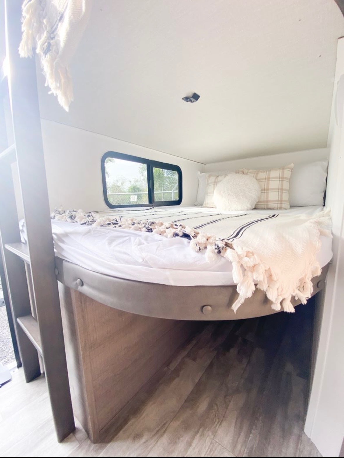 Cozy RV rental sleeping area featuring an elevated bed, soft bedding, decorative pillows, and a window view.. Grand Design Imagine 2020