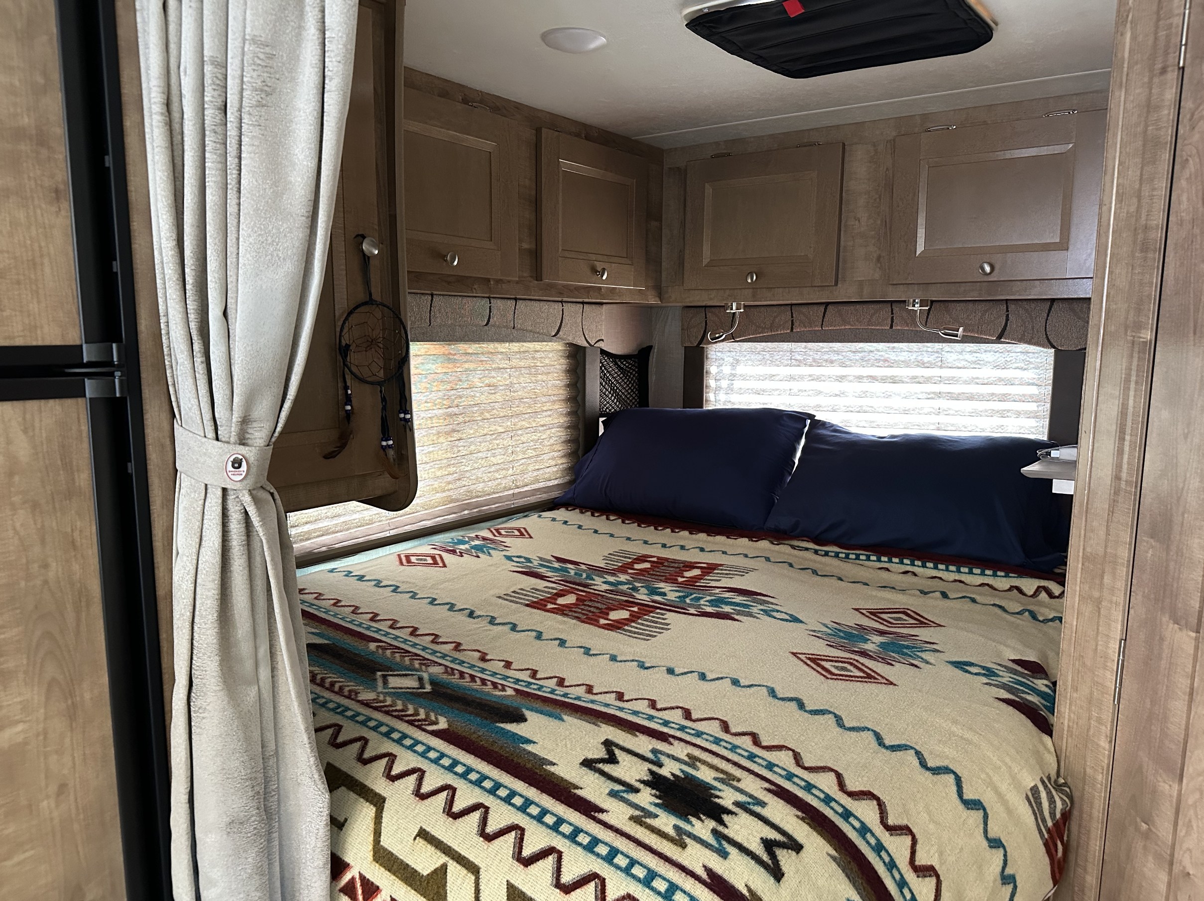 Cozy RV rental bedroom featuring a bed with a southwestern-style blanket, blue pillows, and overhead wooden cabinets.. FORD COACHMAN LEPRECHAUN 2020