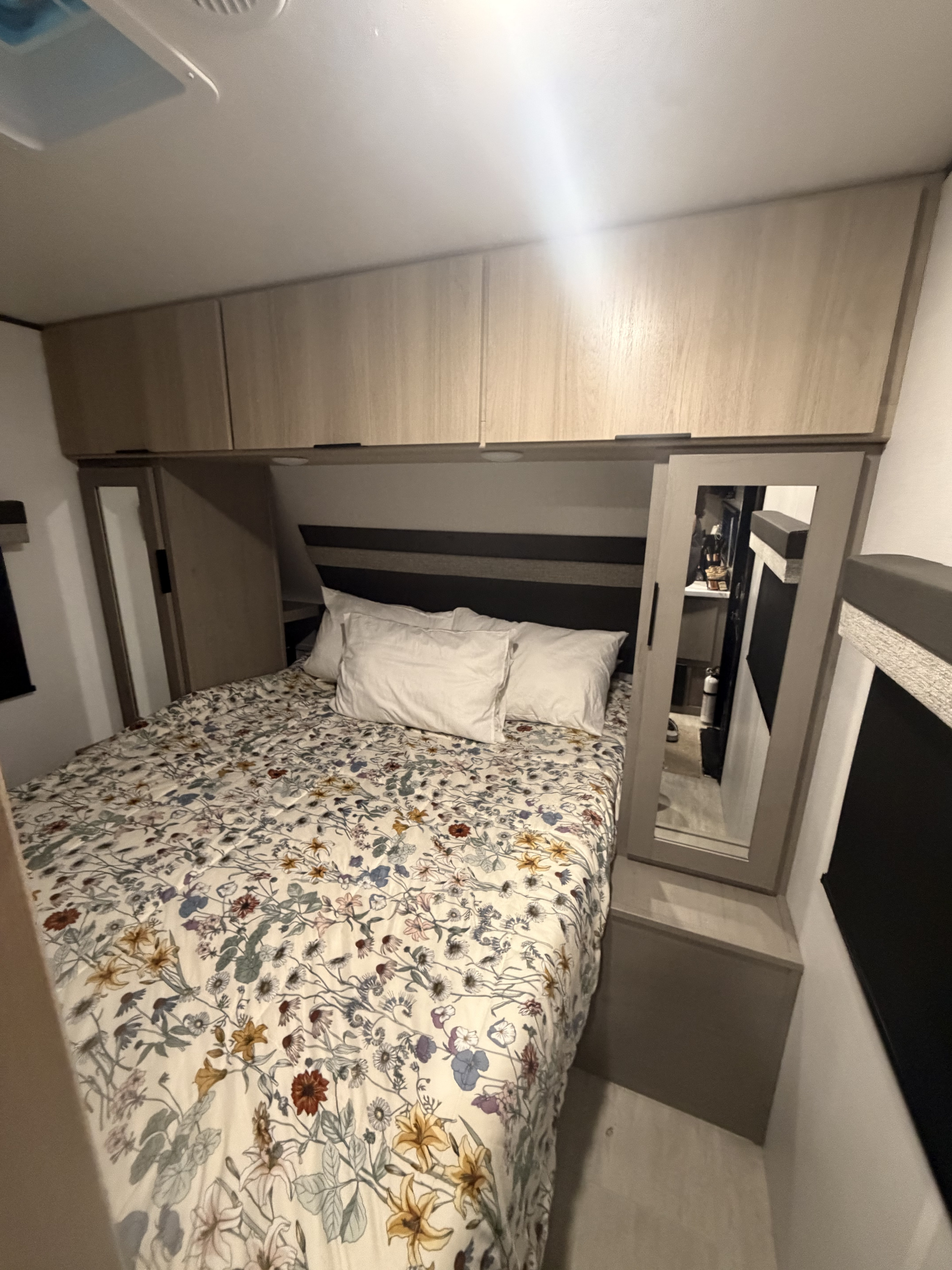 Compact RV rental bedroom featuring a floral bed, overhead storage cabinets, and a mirrored wardrobe for practical travel.. Dutchmen Colorado 2025