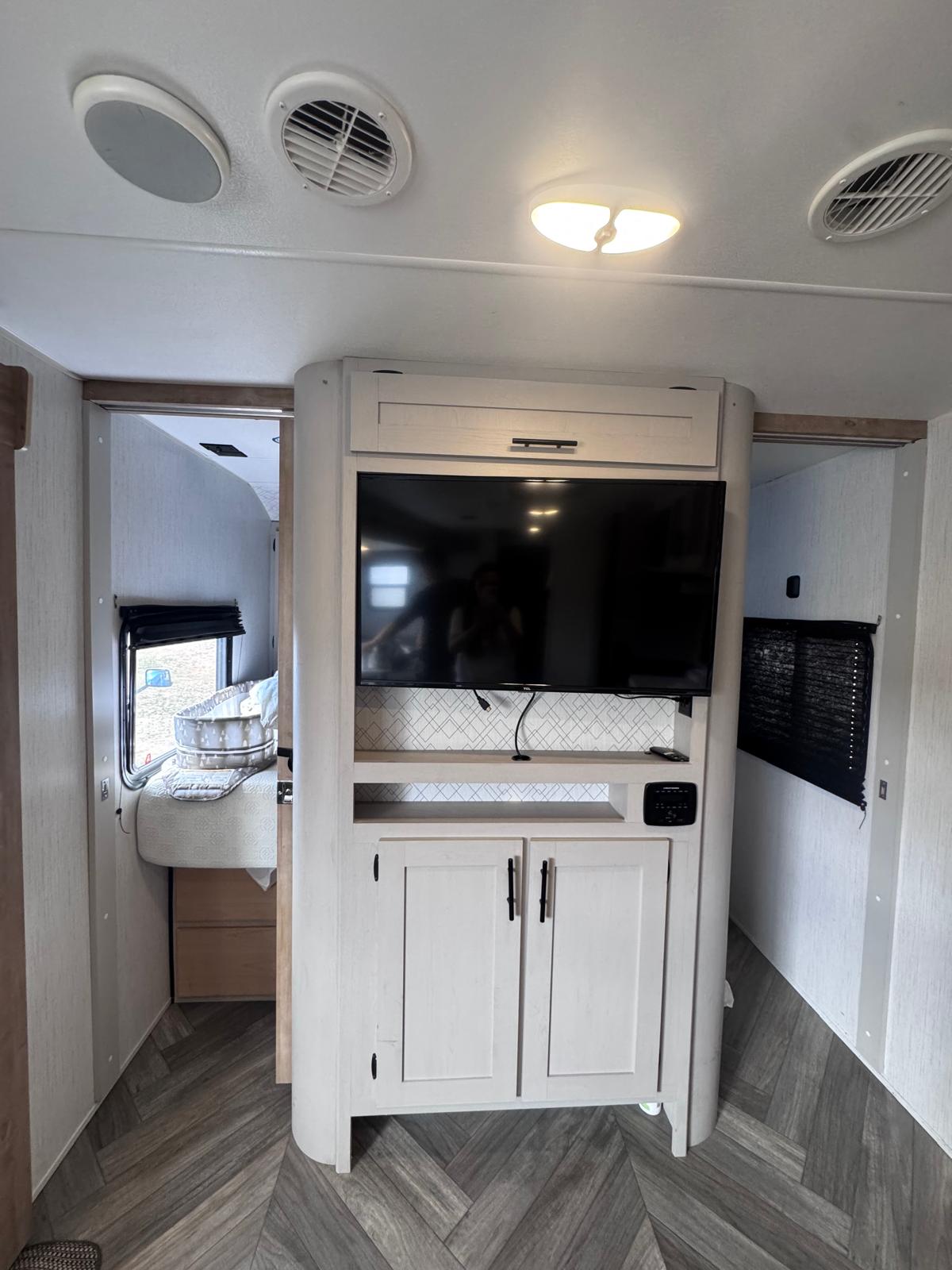 Modern RV rental interior with a central TV entertainment unit, light wood cabinets, and herringbone flooring, separating two living areas.. Heartland RVs Prowler 2023