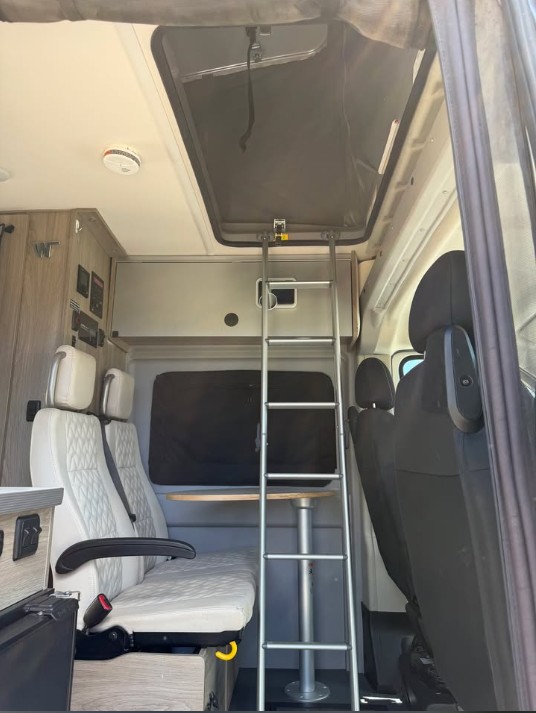 RV rental camper van interior. Ladder to upper bed, with white quilted seats and a small table below.. Winnebago Solis 2022