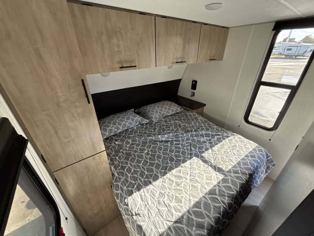 Experience comfort in this RV rental bedroom: a bed with patterned bedding, overhead cabinets, and a window for views.. Forest River Grey Wolf 2026