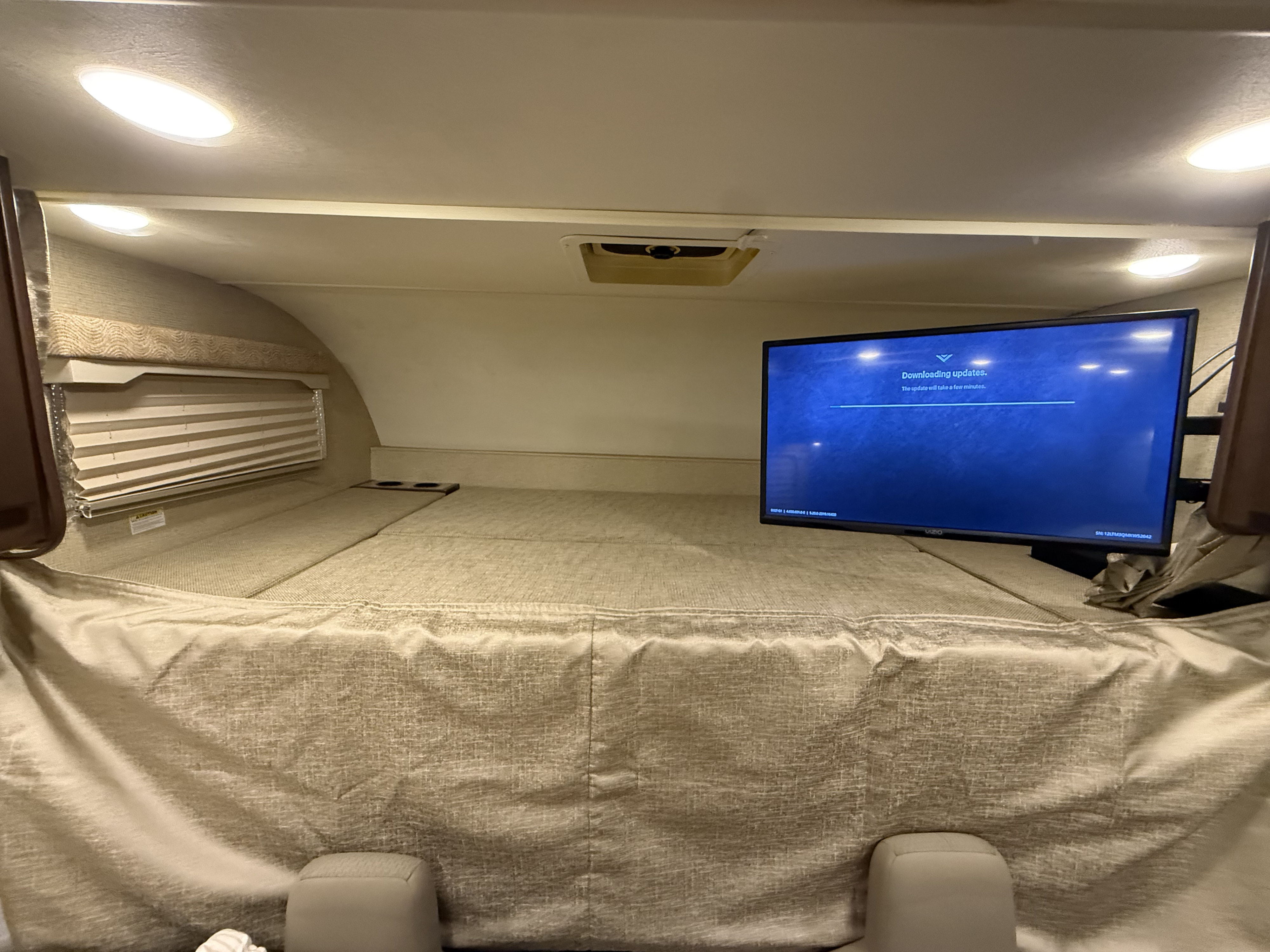 Inside an RV rental, a beige cab-over bunk bed with a TV on the right showing "Downloading updates." Overhead lights glow.. Thor Freedom Elite 2017