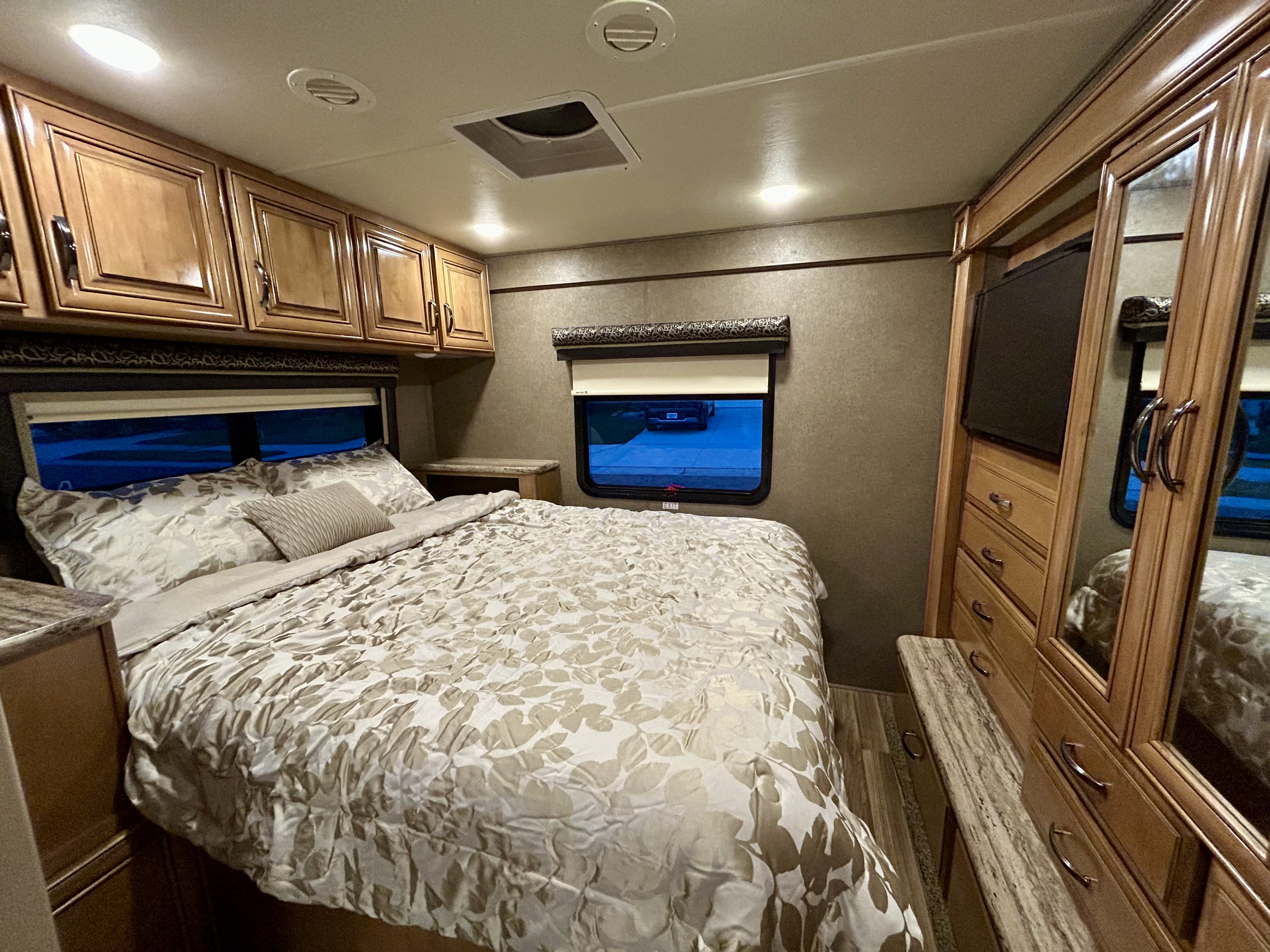 Cozy RV rental bedroom featuring a patterned bed, rich wood cabinets, and mirrored storage, ideal for comfortable travel.. Thor Quantum 2017