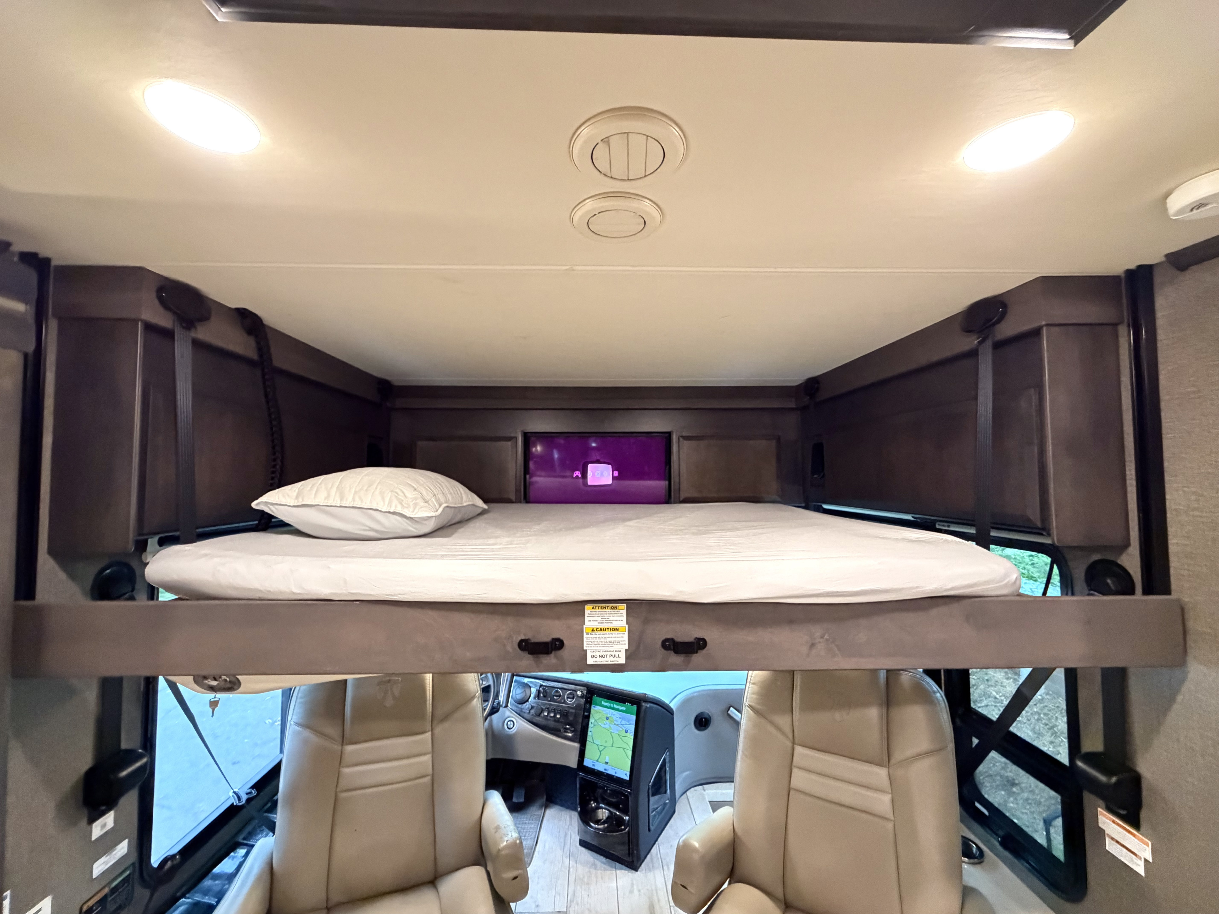Overhead bunk bed with a TV in this RV rental, overlooking the driver's cockpit with its dashboard and tan seats.. Thor Venitian 2021