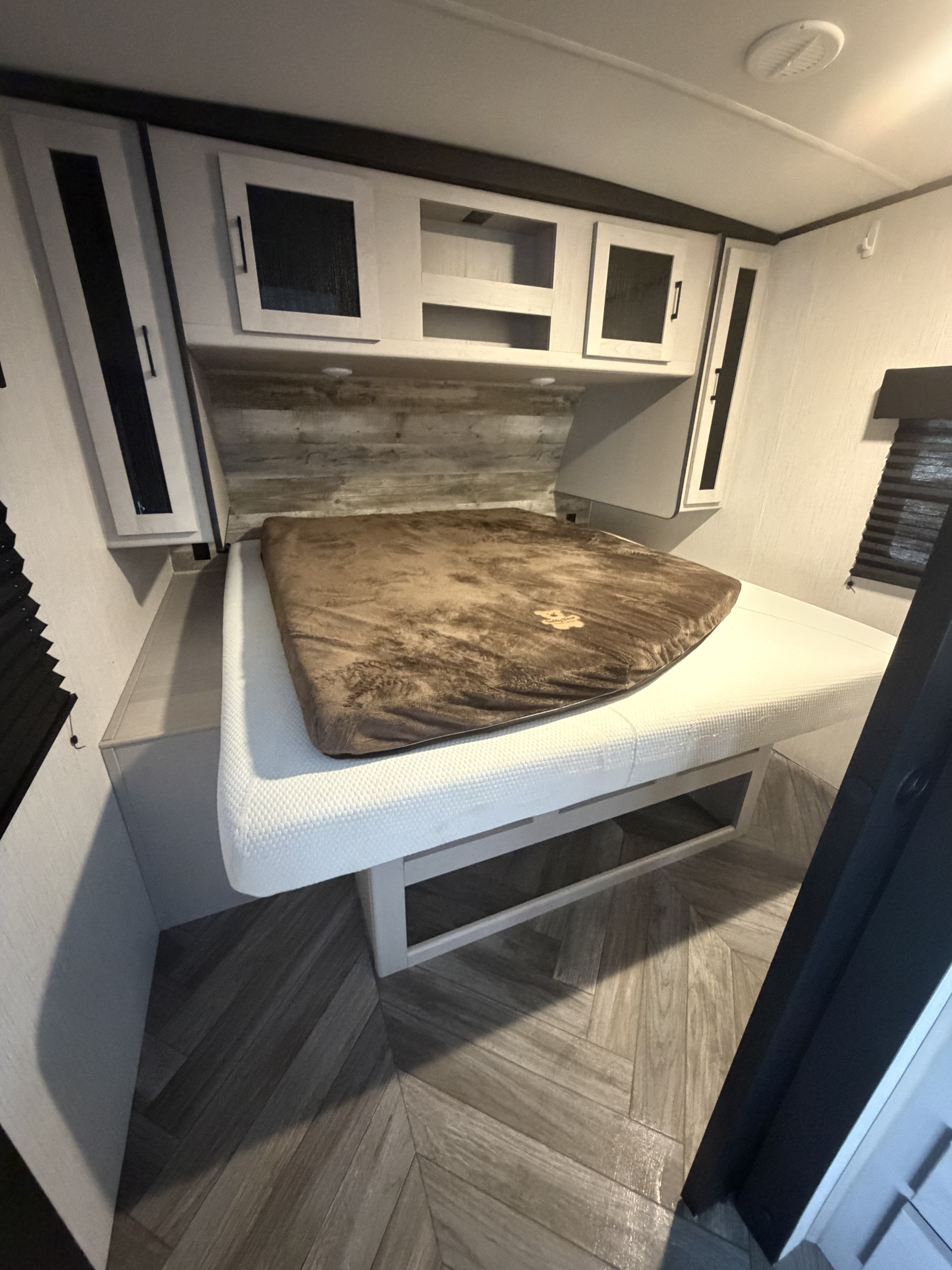 This RV rental bedroom has a cozy bed with a brown topper, overhead cabinets, and herringbone flooring.. Heartland RVs Mallard 2023