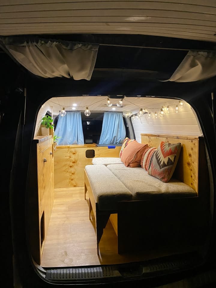 Inside a cozy RV rental van conversion, featuring comfy seating, wood interior, string lights, and storage.. Hyundai Kickback Camper 2011
