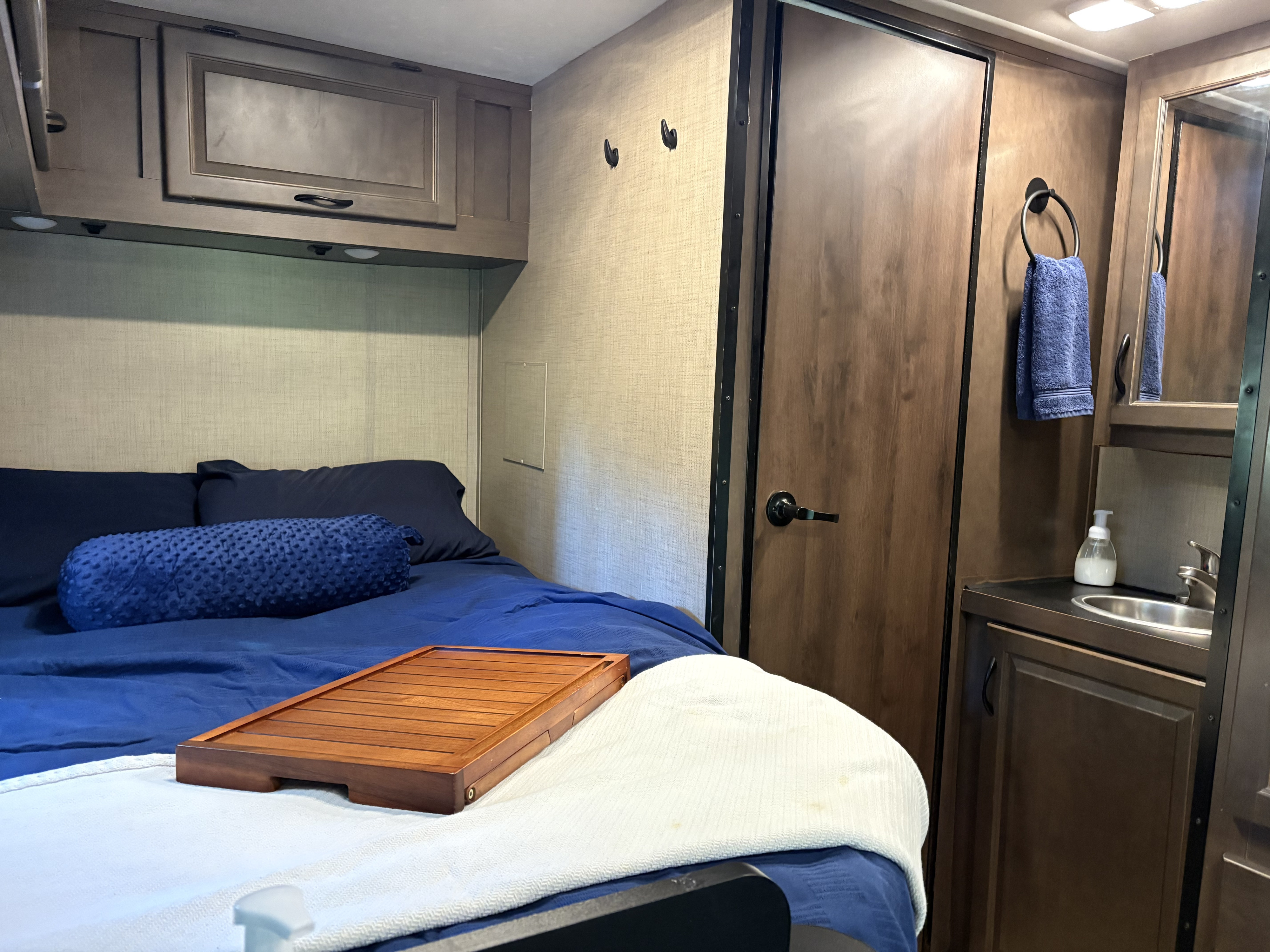 Experience comfort in this rv rental. View of a cozy bedroom with blue bedding, overhead cabinets, and an adjacent bathroom with a sink.. Wayfarer Adventure 2020