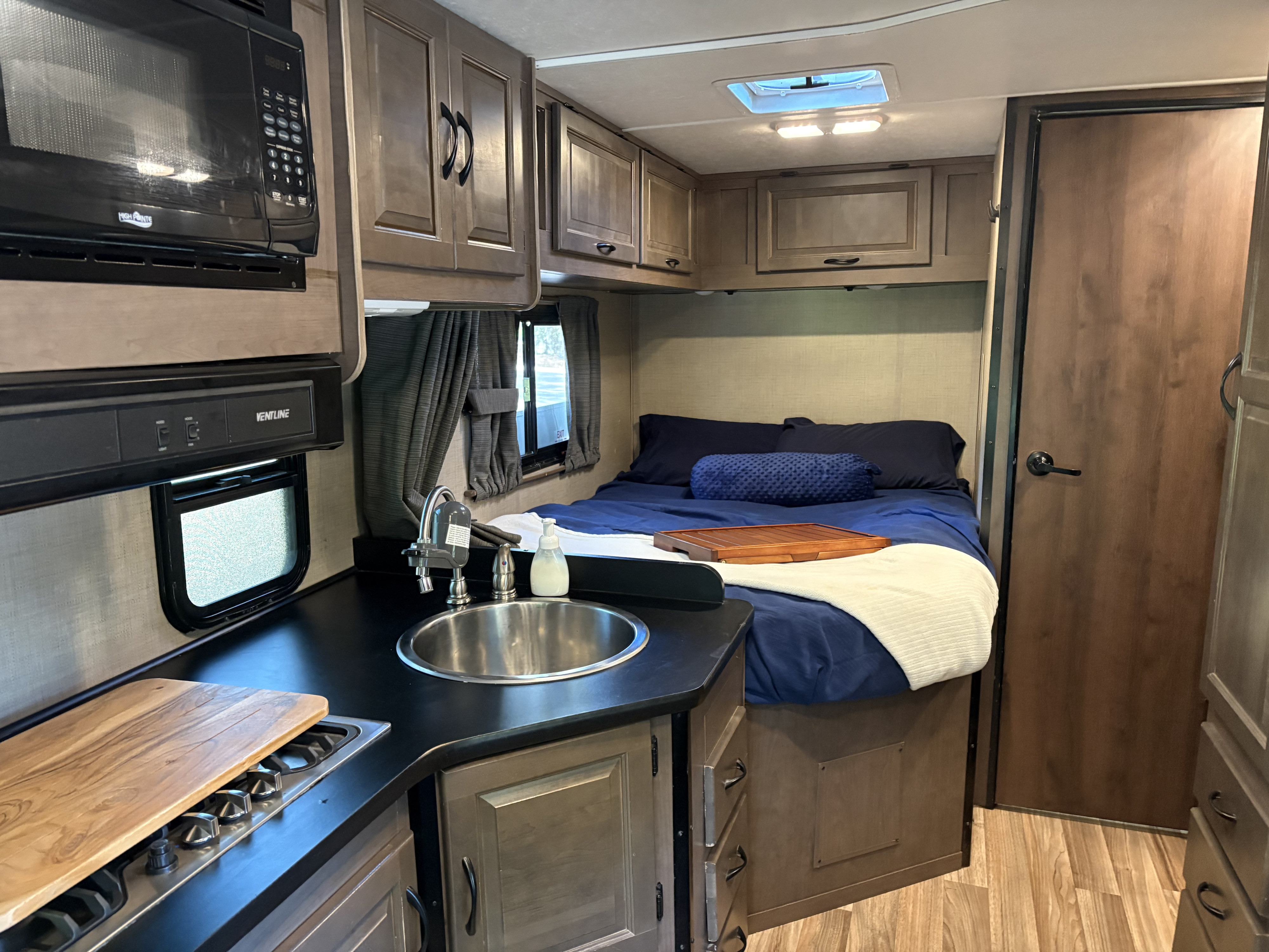 A cozy RV rental interior featuring a kitchen with microwave, sink, and stove, alongside a comfortable bed with blue bedding.. Wayfarer Adventure 2020