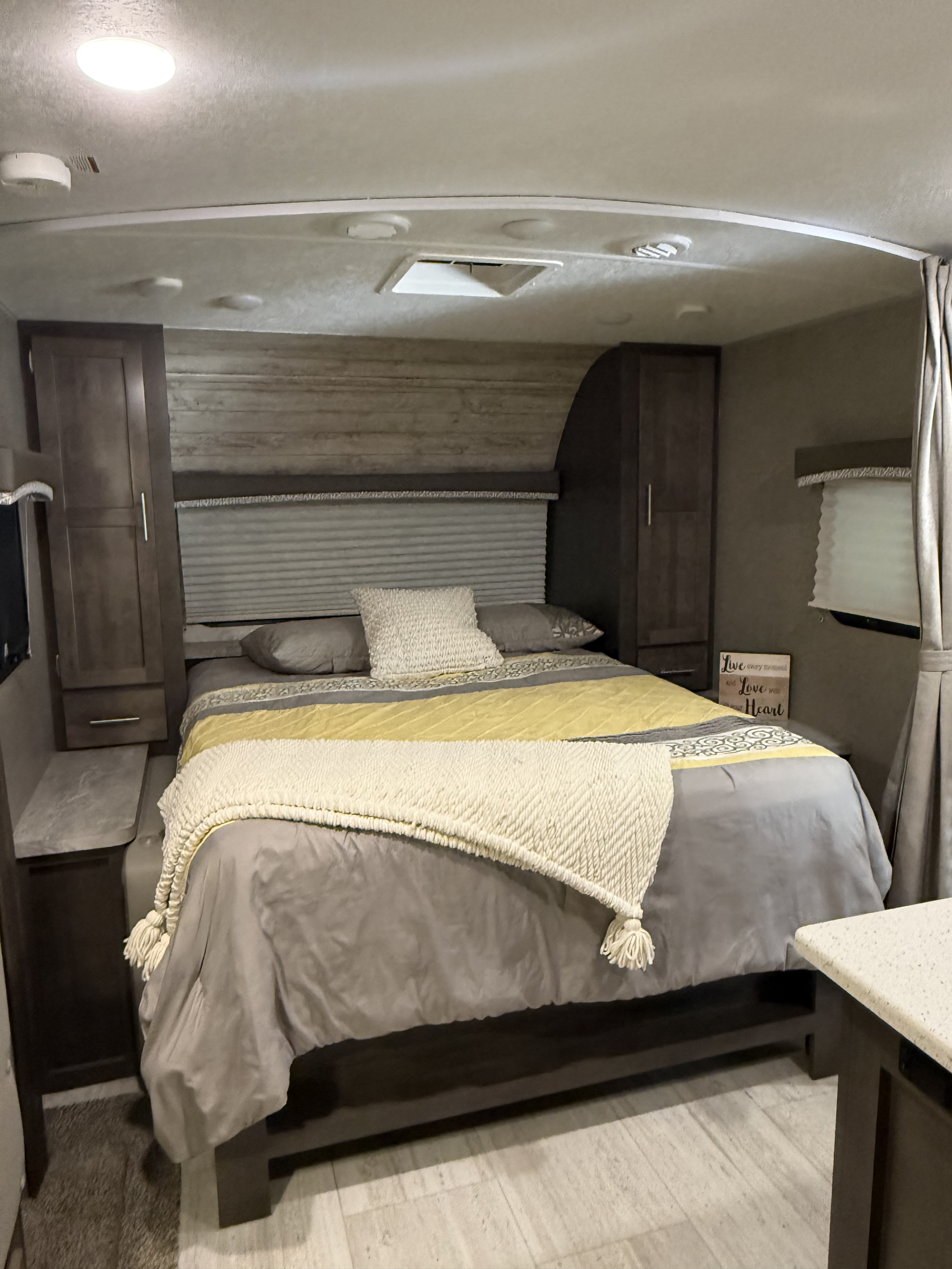 This cozy RV rental bedroom features a bed with gray & yellow bedding, white throw, wood cabinets, and light flooring.. Forest River Rockwood Mini Lite 2019