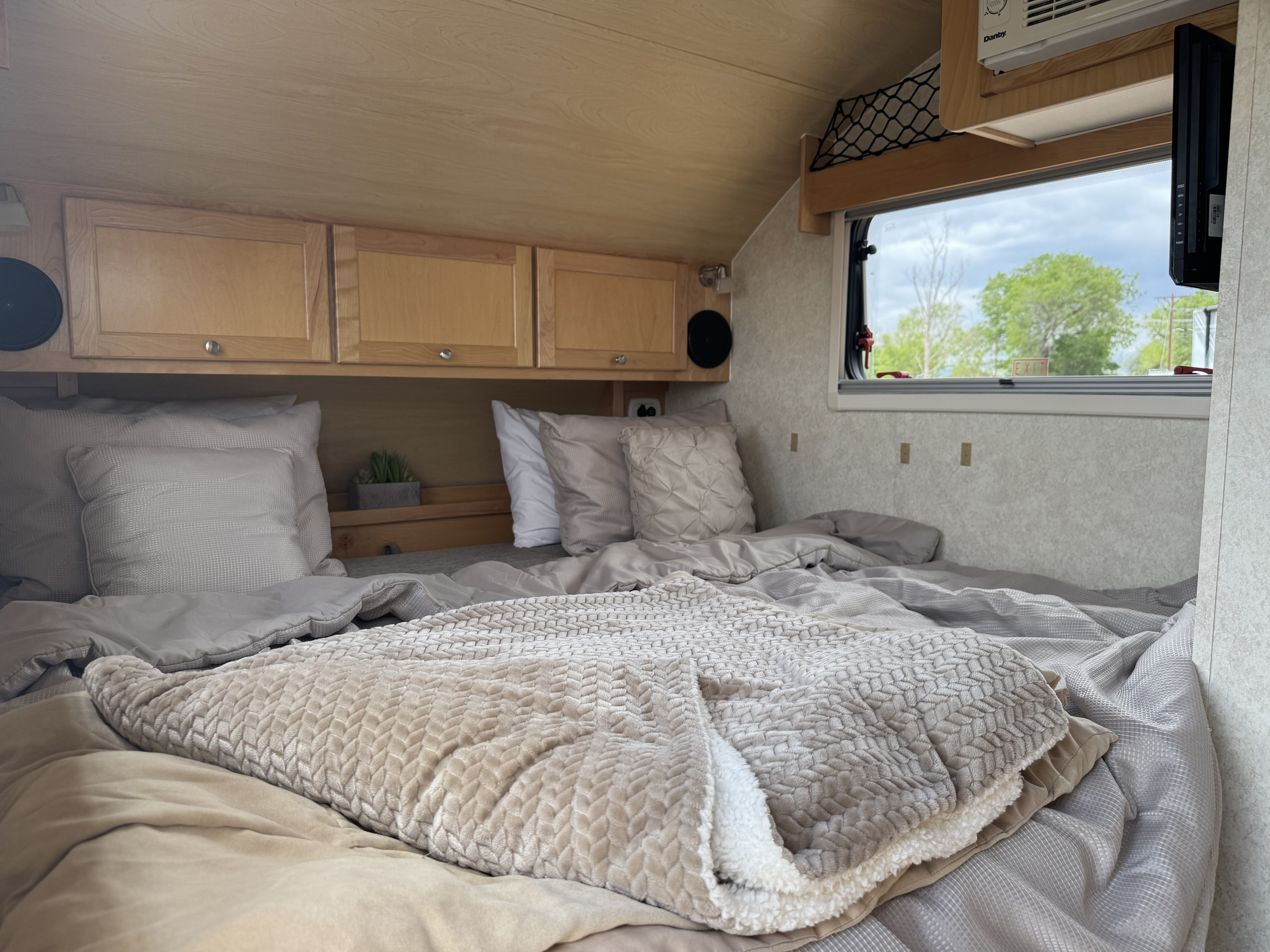 Inviting RV rental interior: a plush bed with neutral pillows/blankets, wood cabinets, and window view for a cozy camping trip.. nuCamp T@B 2018