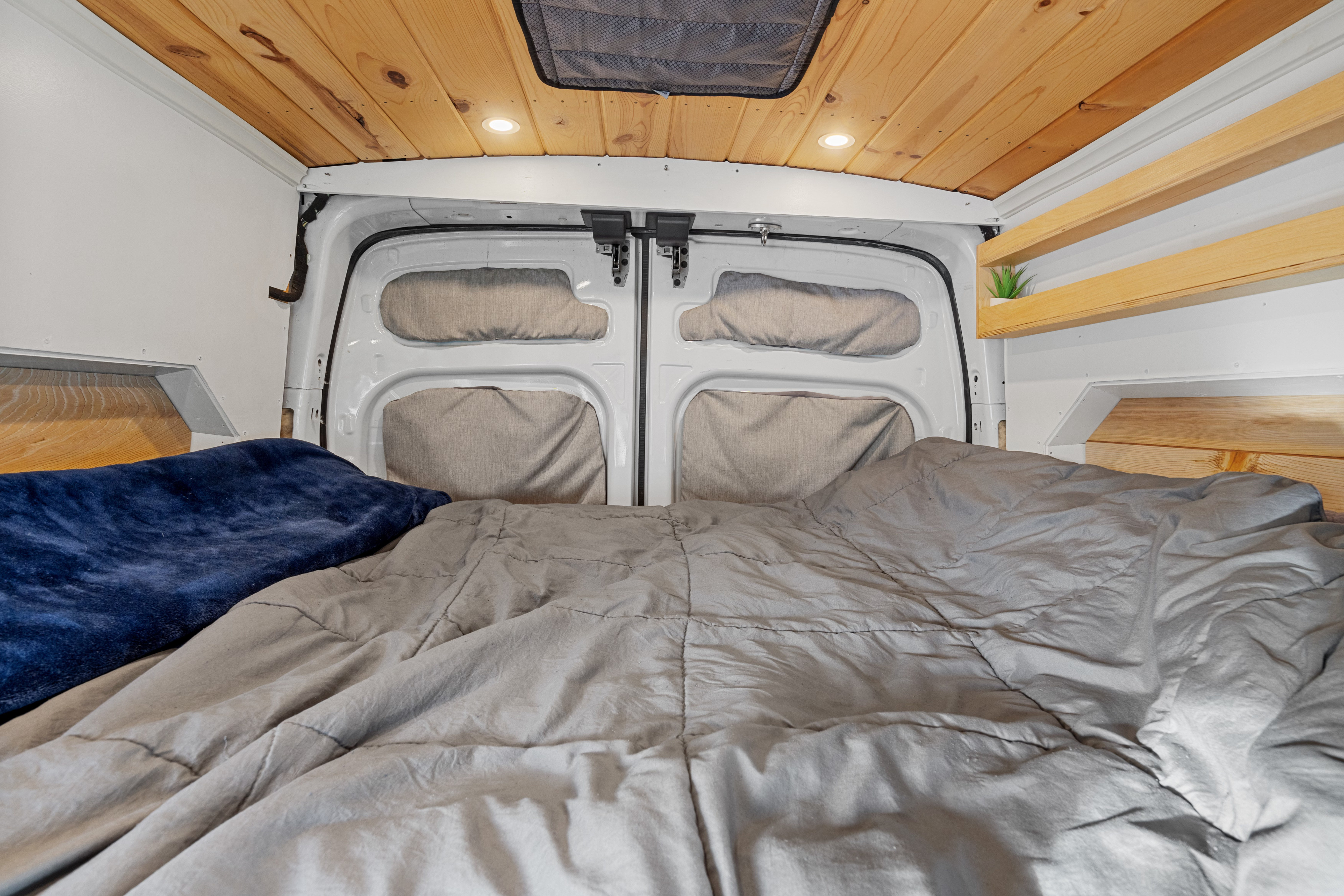 Cozy RV rental bed area with gray comforter, blue blanket, wooden plank ceiling, recessed lights, and rear van doors.. Ford Transit Custom 2017
