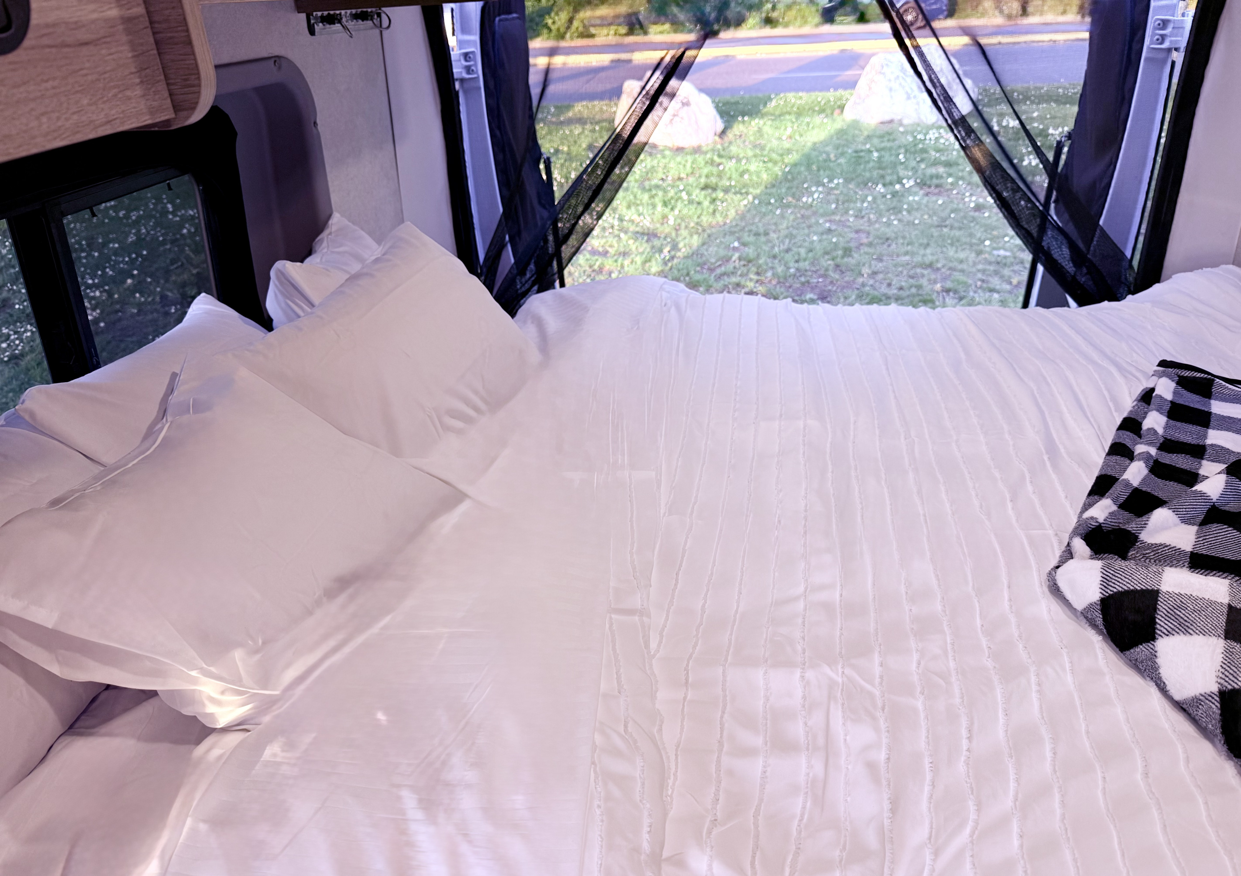 Cozy bed in an RV rental with white sheets, a buffalo check blanket, and a view of nature through screened windows.. Winnebago Solis 2025