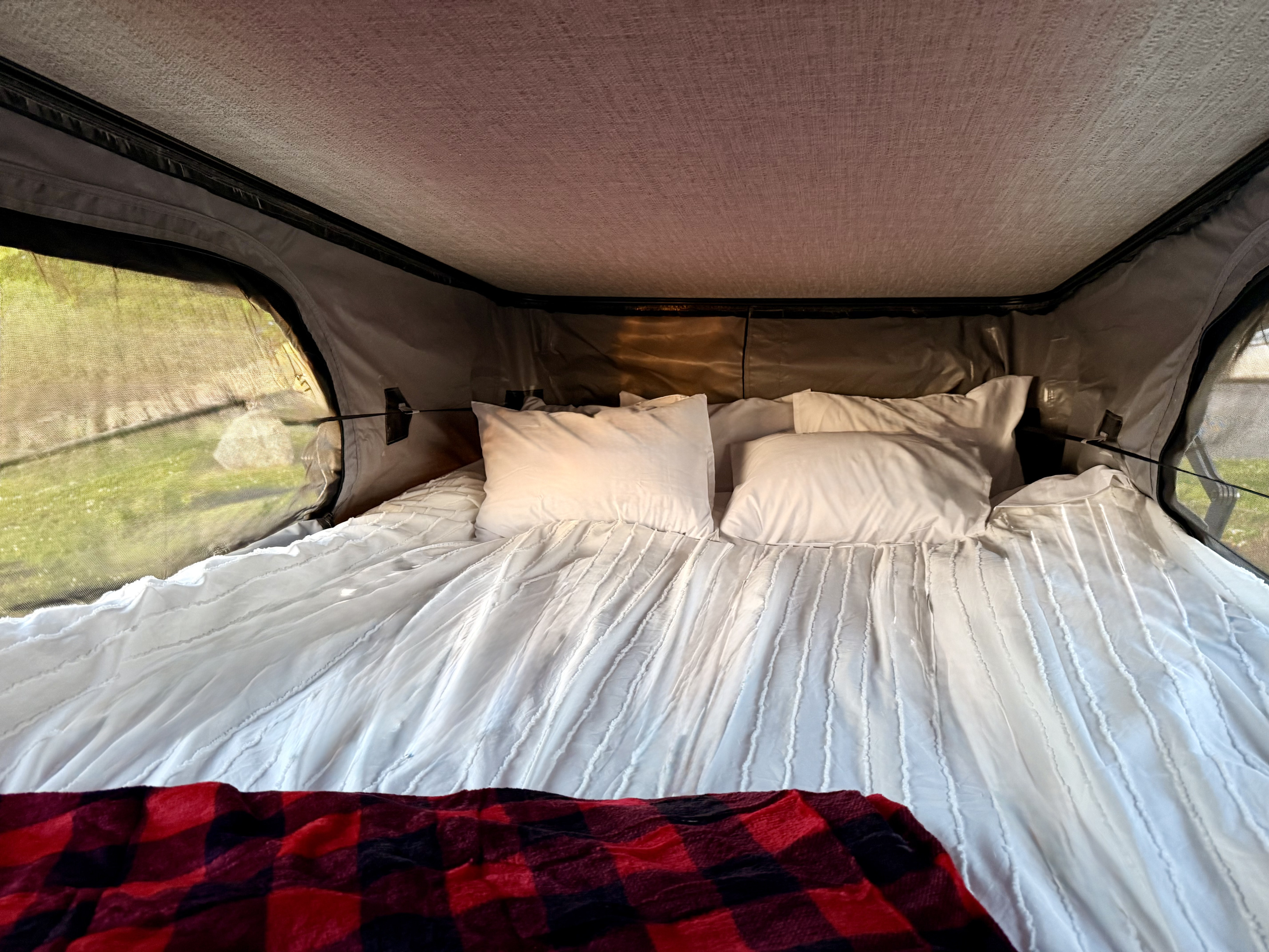 A comfy bed with white bedding & a red plaid blanket inside an RV rental tent, offering views of green outdoors.. Winnebago Solis 2025