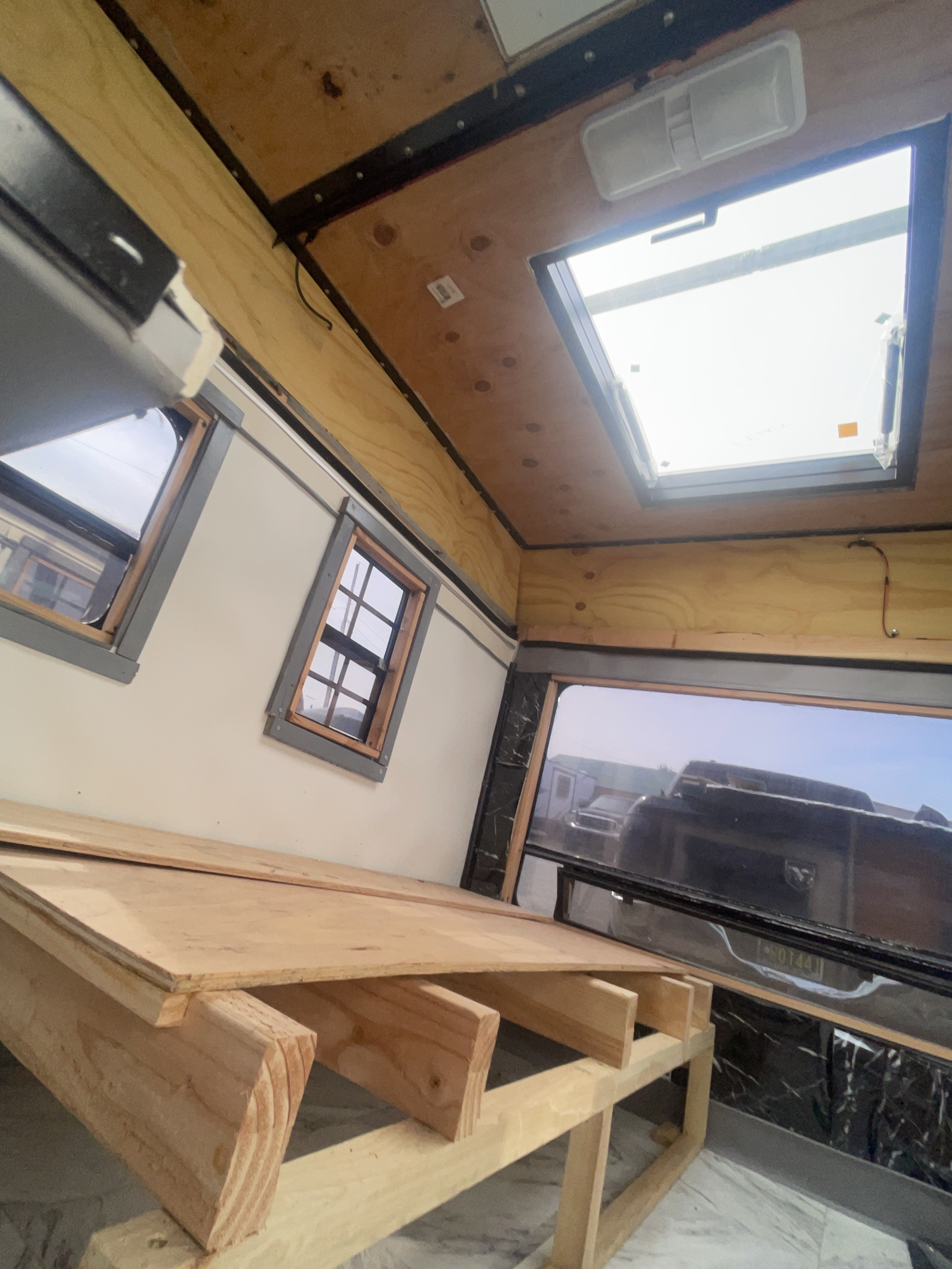RV rental interior under construction, featuring a wooden bed platform, bright skylight, and framed windows.. Camping Equipment Cargo Trailer Conversion 2020