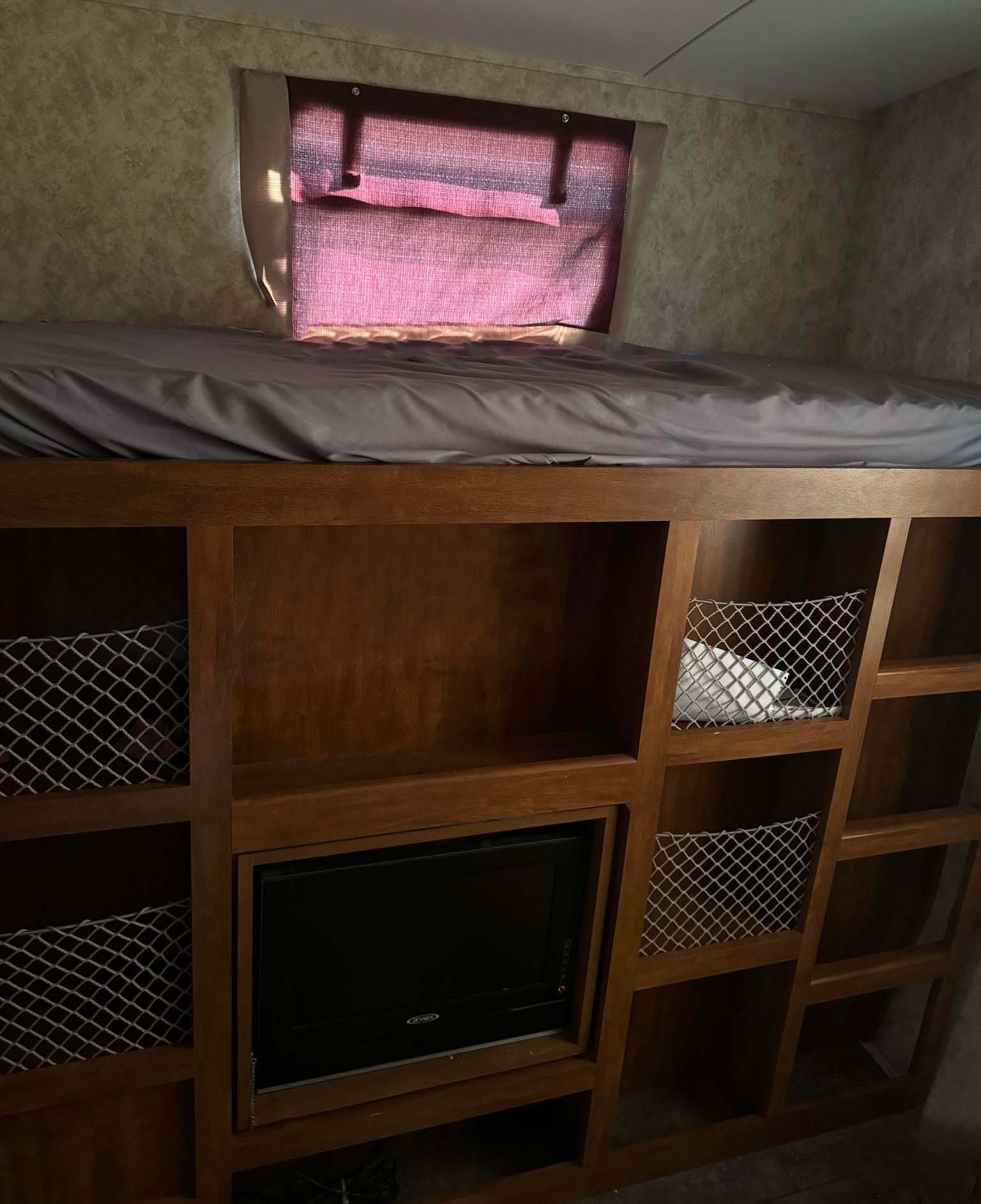 RV rental bunk bed area: a top bunk with grey sheets, wooden storage shelves with mesh nets, and a small TV below.. Keystone RV Bullet Premier Ultra 2011