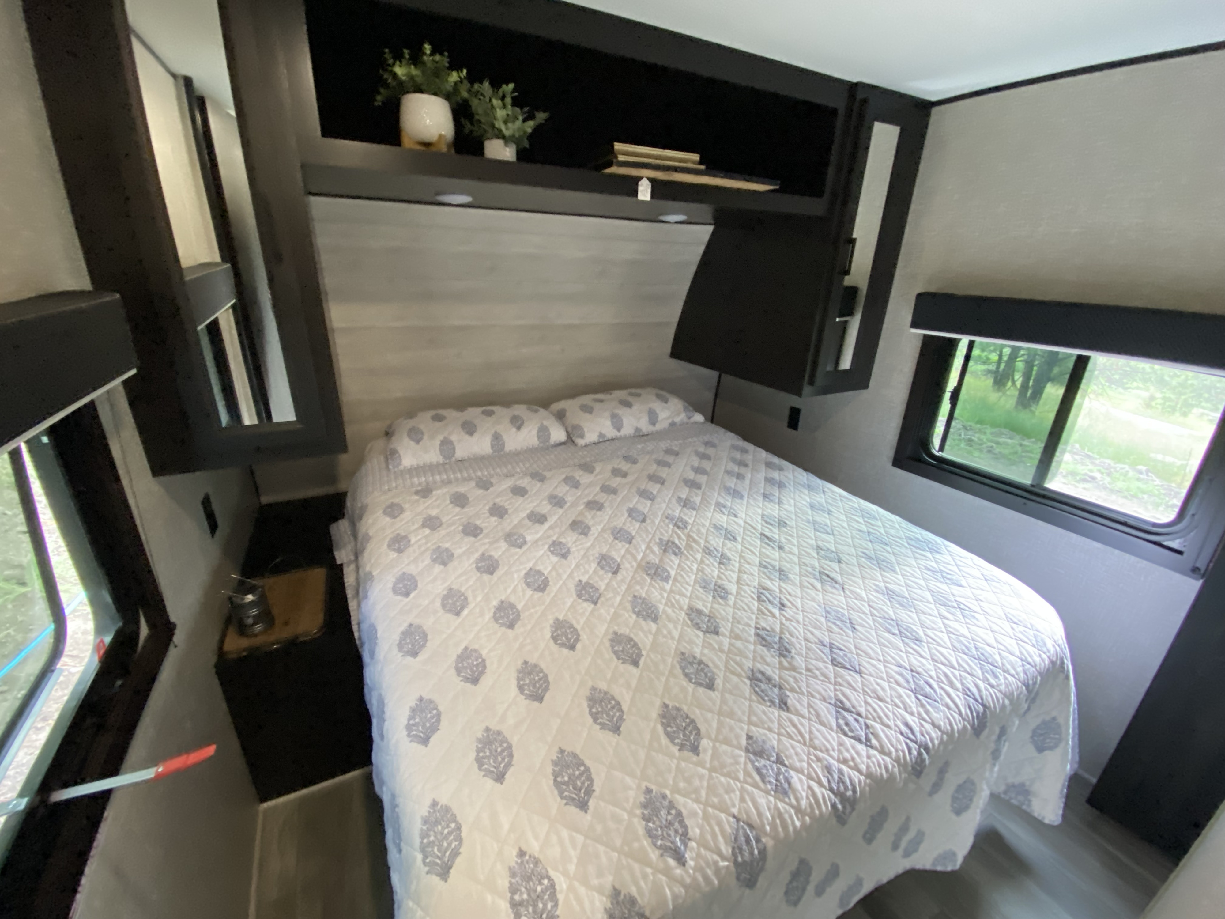 Cozy RV rental bedroom with patterned white and gray bedding, overhead storage, and windows revealing lush greenery outside.. Jayco Jay Flight 2023