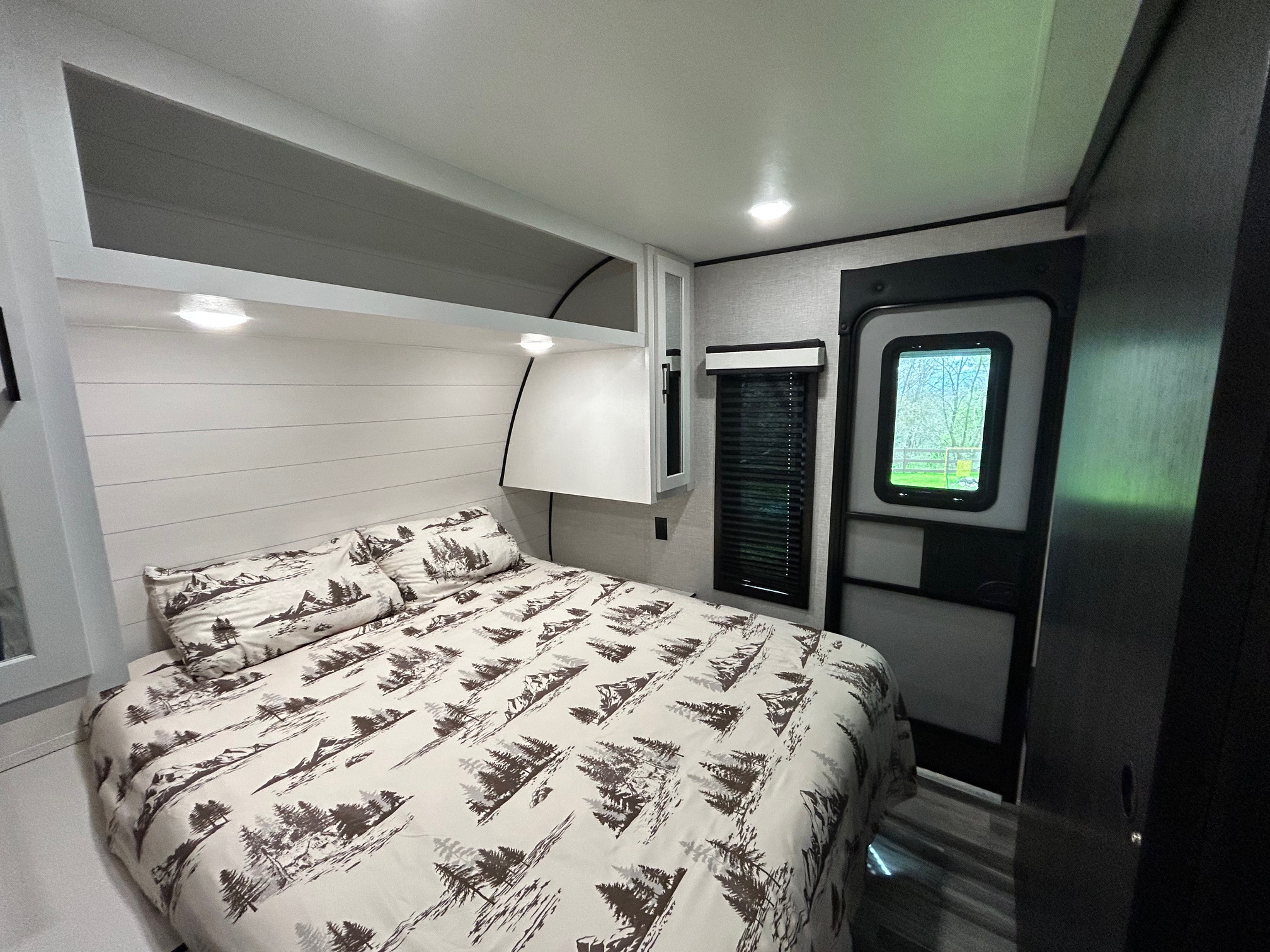 A compact yet inviting RV rental bedroom features a bed with scenic tree bedding, upper cabinets, reading lights, and an entry door.. Jayco Jay Flight 2024