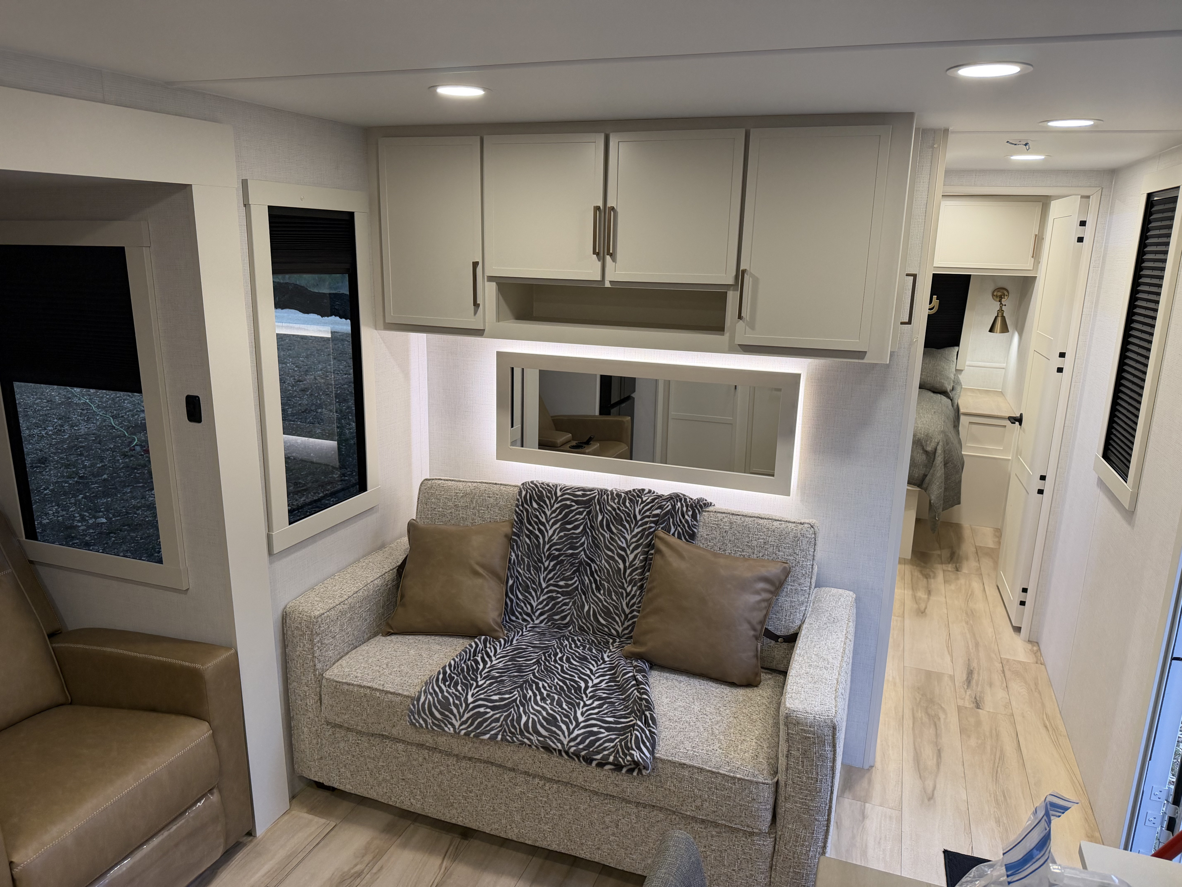 Modern rv rental living area features a grey couch with accent pillows, overhead cabinets, and a hallway leading to the bedroom.. Brinkley RV Model I 2025