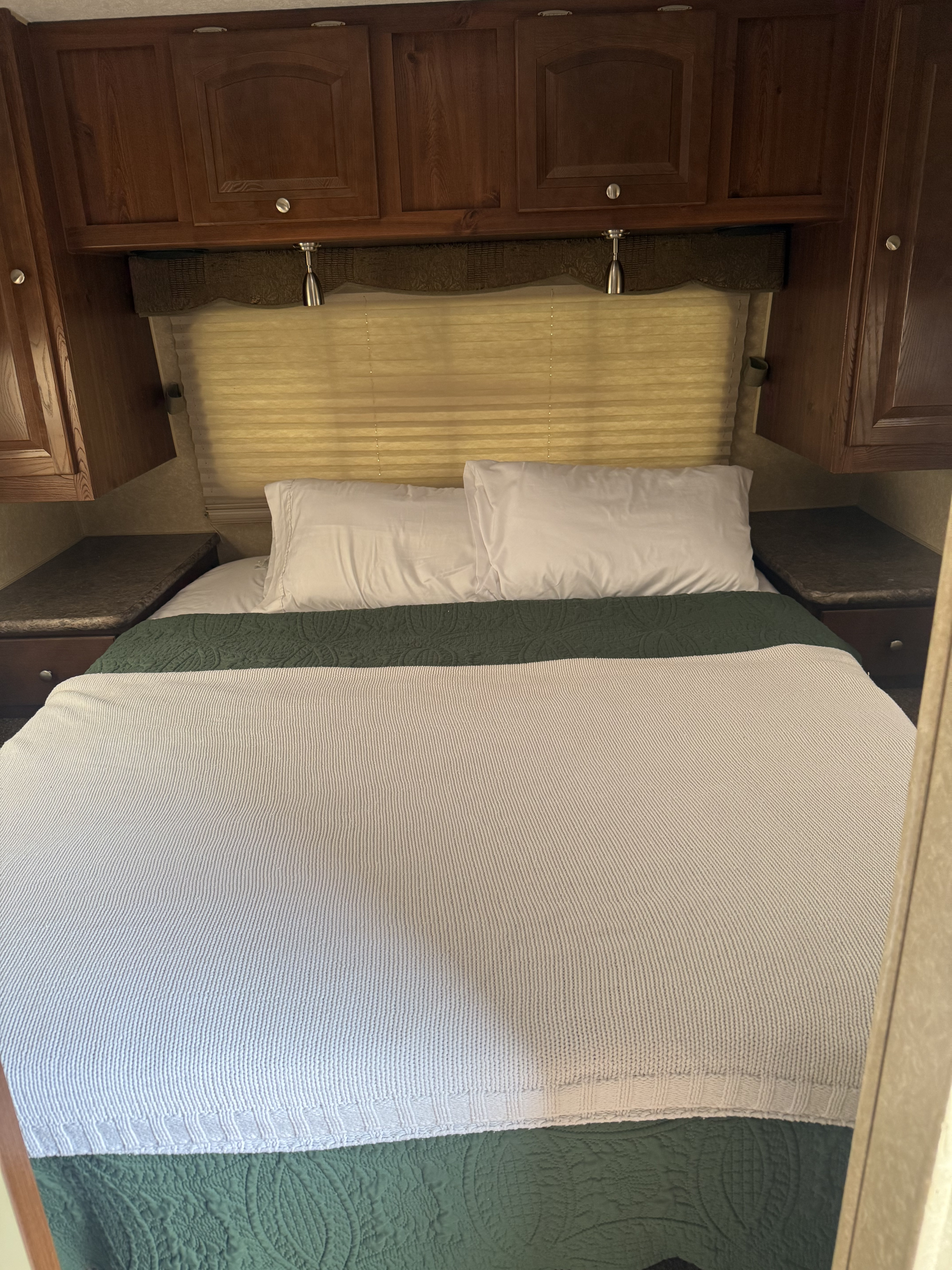 Cozy RV rental bedroom features a bed with white pillows, green quilted blanket, overhead wood cabinets, and window blinds.. Forest River Rockwood Signature Ultra Lite 2011