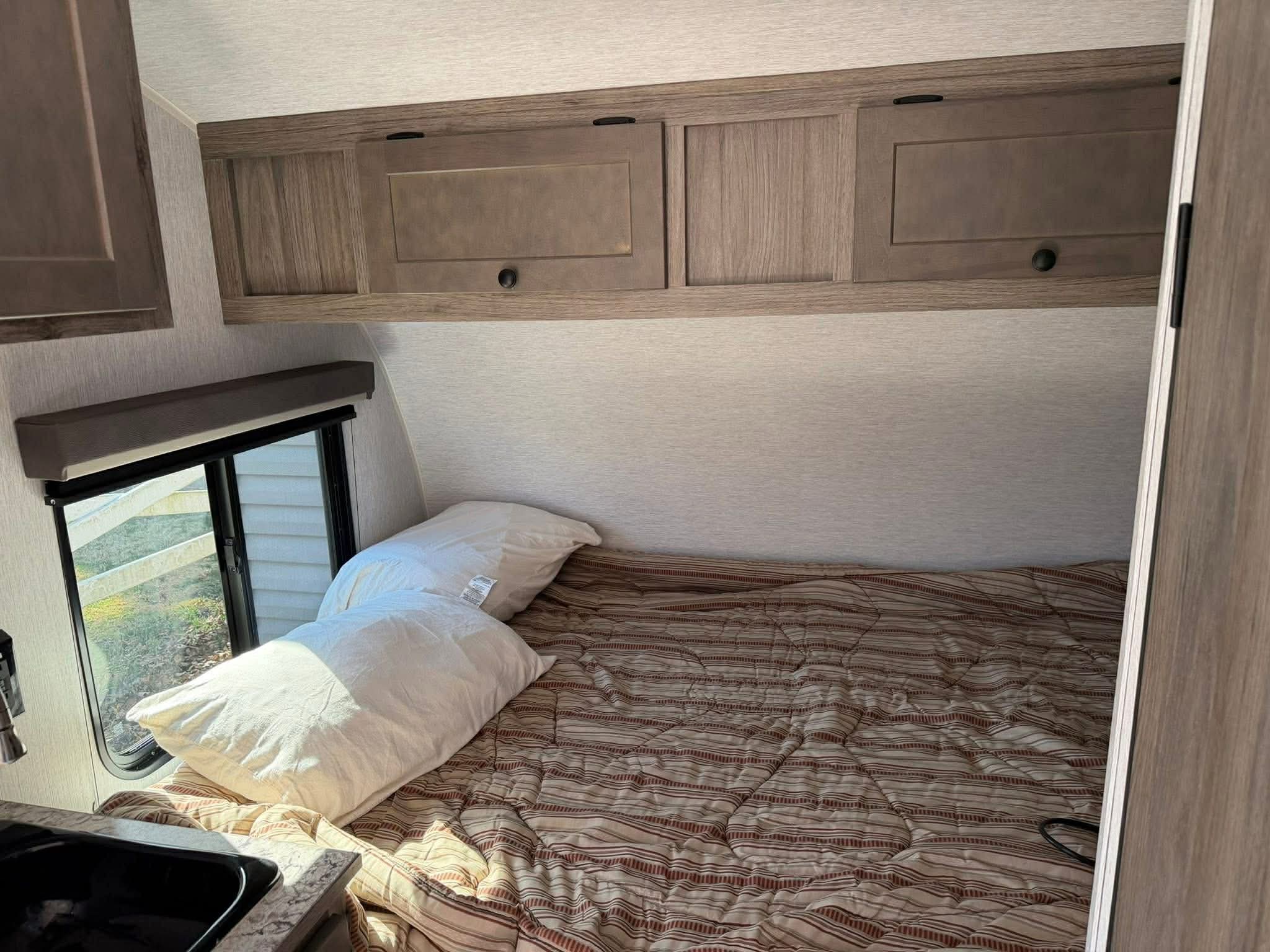 Inside an RV rental: bedroom area with a comfortable bed, wood-look cabinets, and a sunlit window for travelers.. Sunset Park RV SunRay 2025