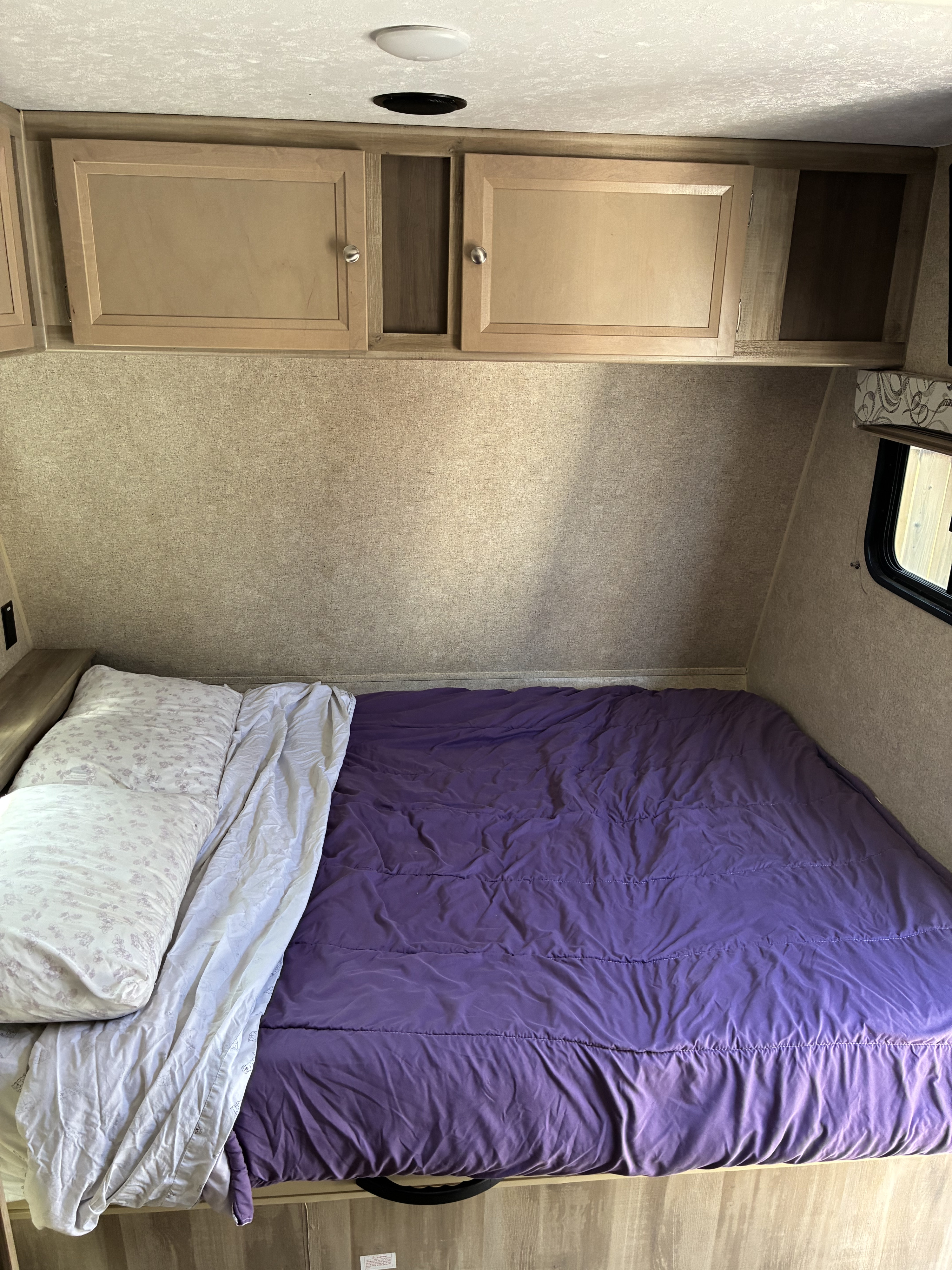 View of a compact RV rental bedroom with a purple bed, white pillow, overhead storage, and light wood accents.. Coachmen Catalina Sumit Series 2019