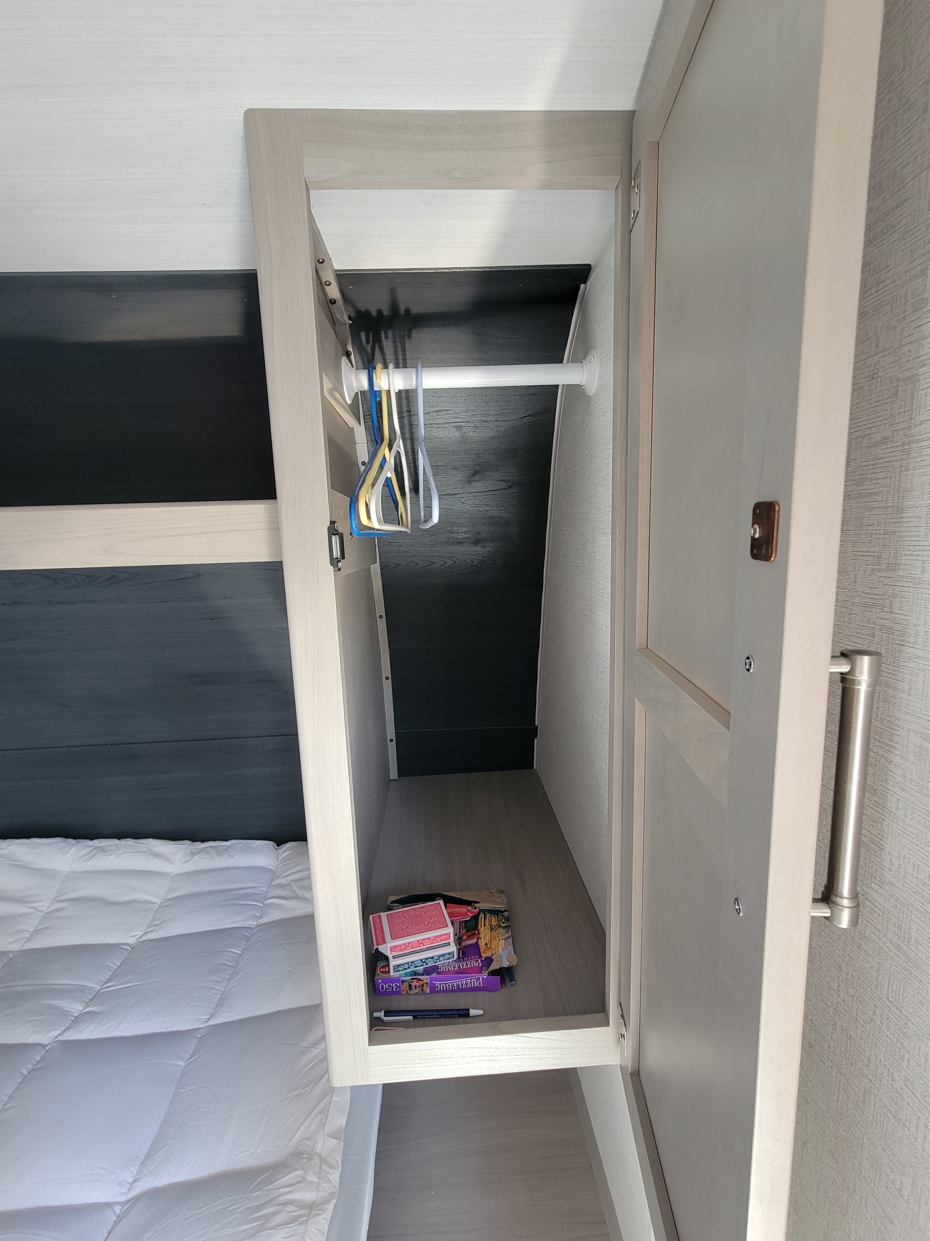 RV rental bedroom with an open closet featuring a hanging rod, hangers, and games on the shelf, next to a white comforter.. Keystone RV Springdale 2024