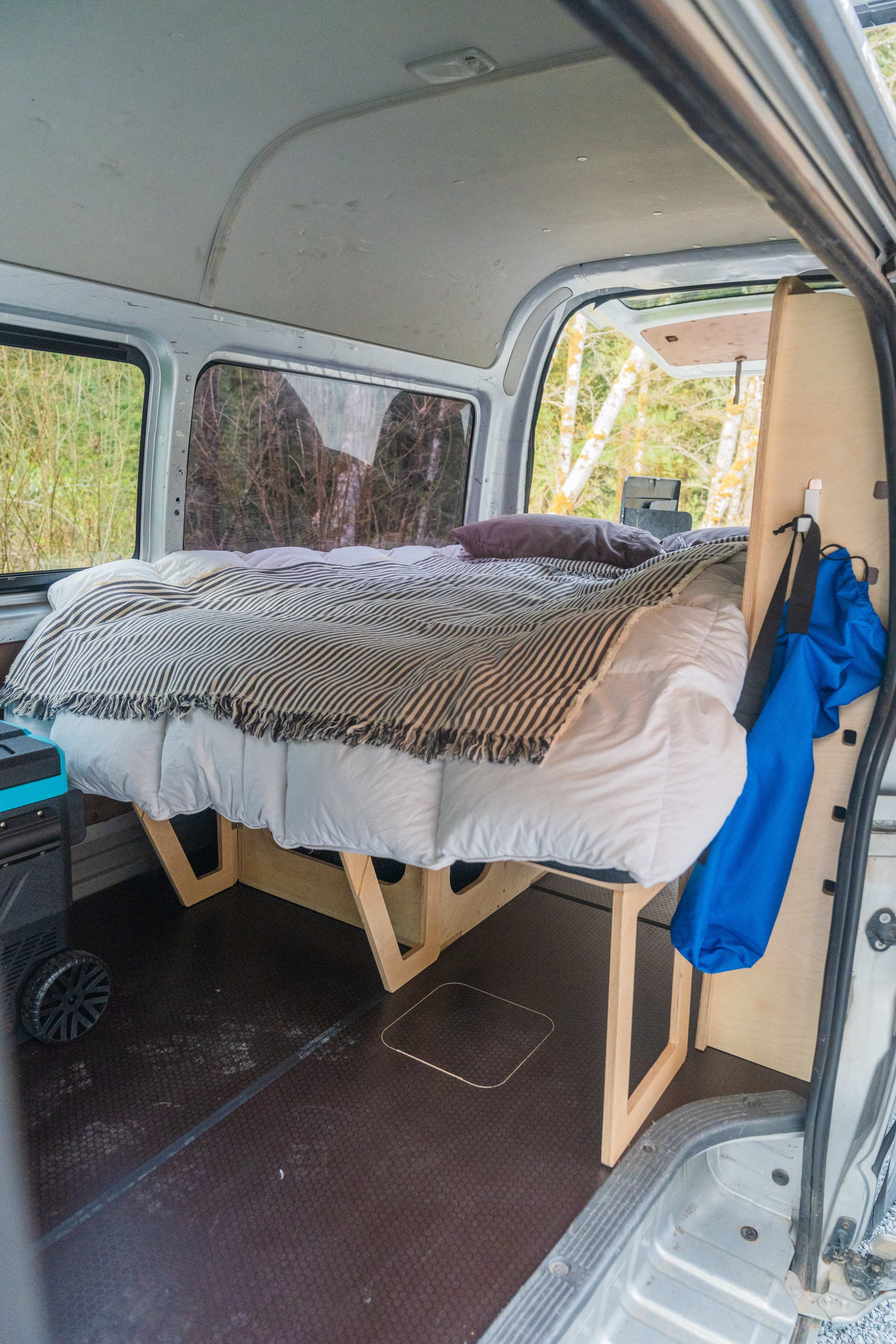 This camper van RV rental features a cozy bed with white bedding, a striped blanket, and a wooden frame overlooking nature.. Toyota Hiace 2005
