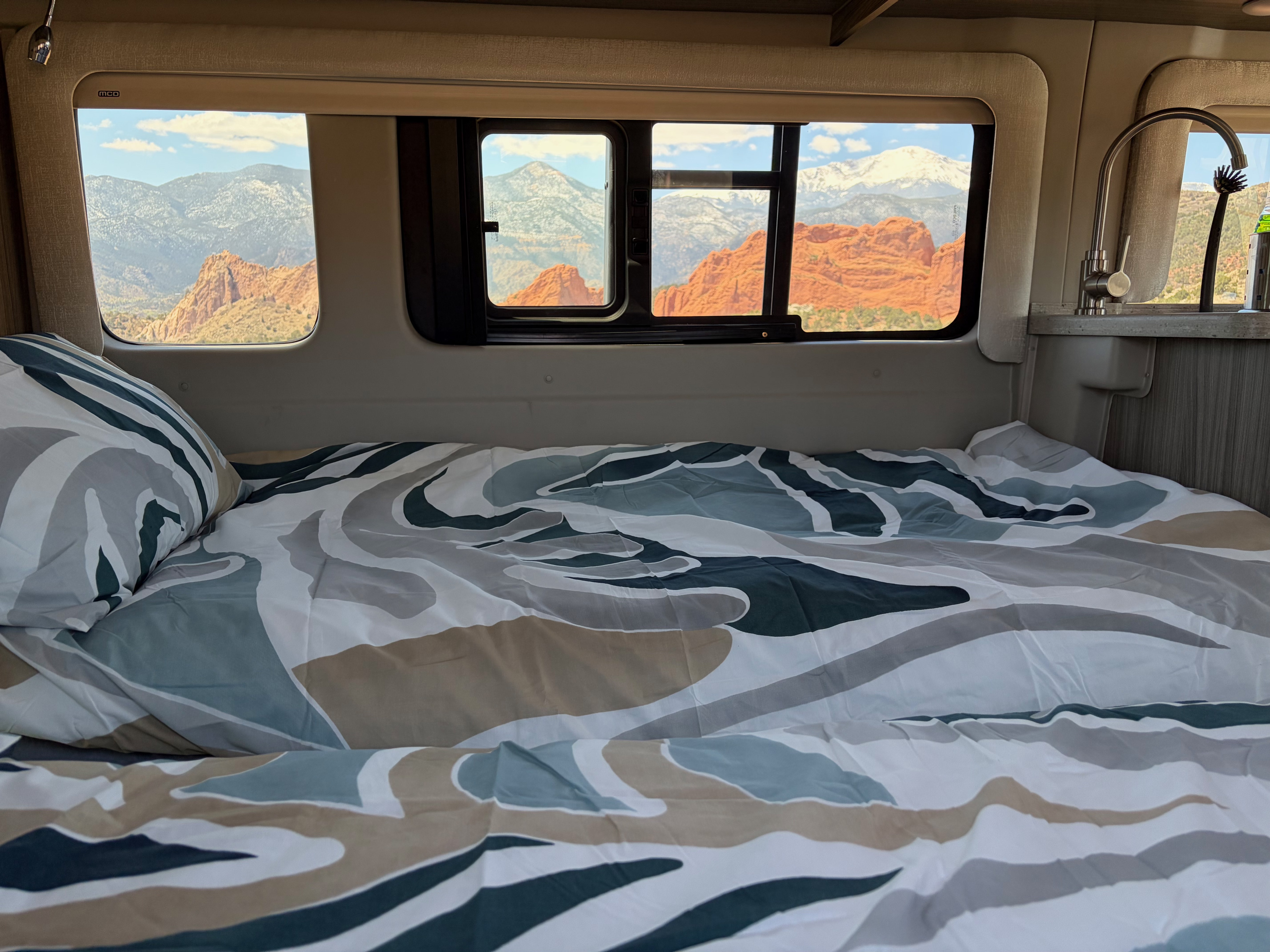 Enjoy breathtaking red rock and snow-capped mountain views from a cozy RV rental bed with patterned bedding and nearby sink.. Entegra Coach Ethos 2023