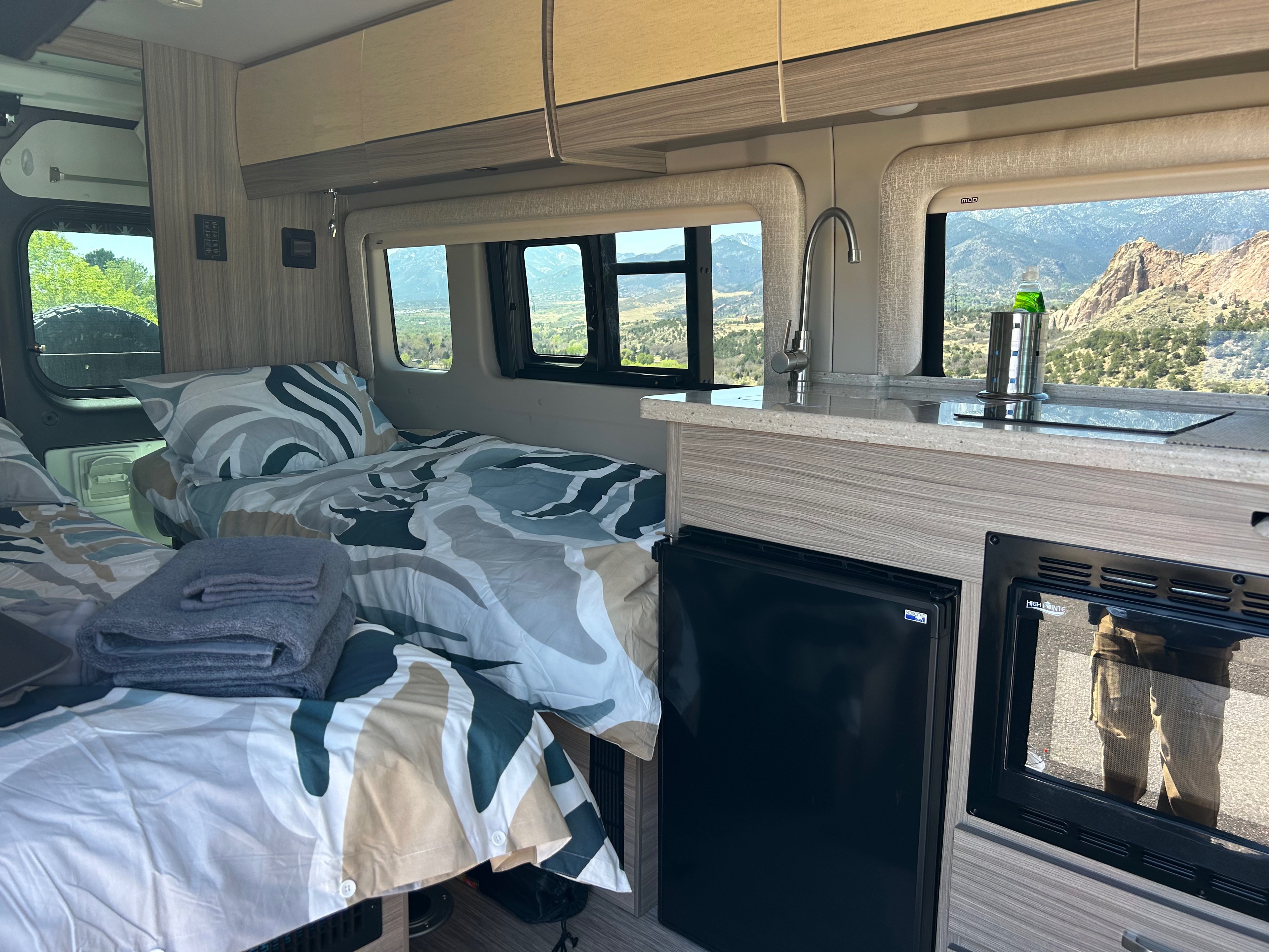 Interior of an RV rental featuring two beds with patterned bedding, a compact kitchen, and scenic mountain views outside.. Entegra Coach Ethos 2023