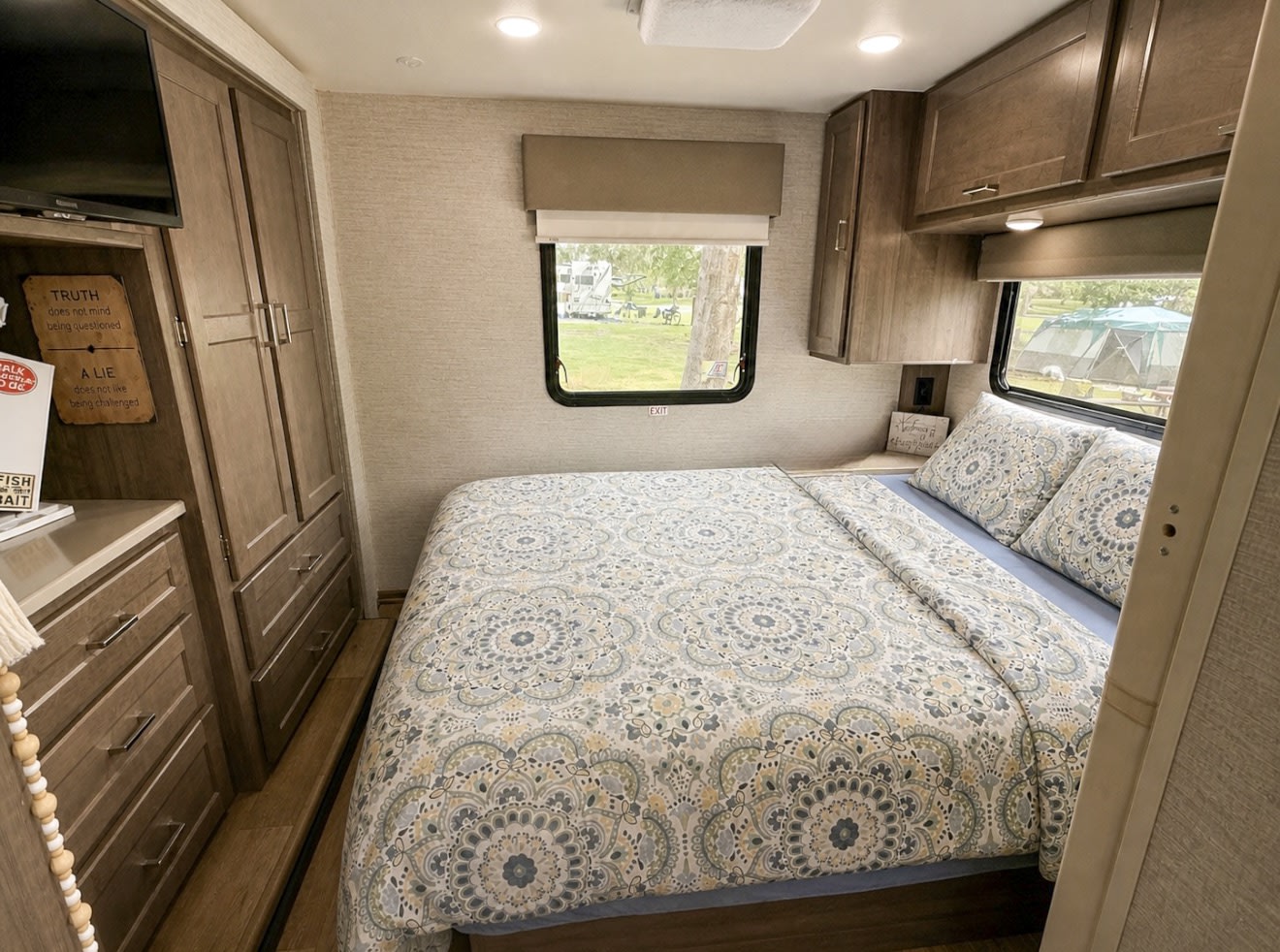 Modern RV rental bedroom with a comfy bed, patterned duvet, wood cabinetry, and windows showing a peaceful campground view.. Winnebago Spirit 2024