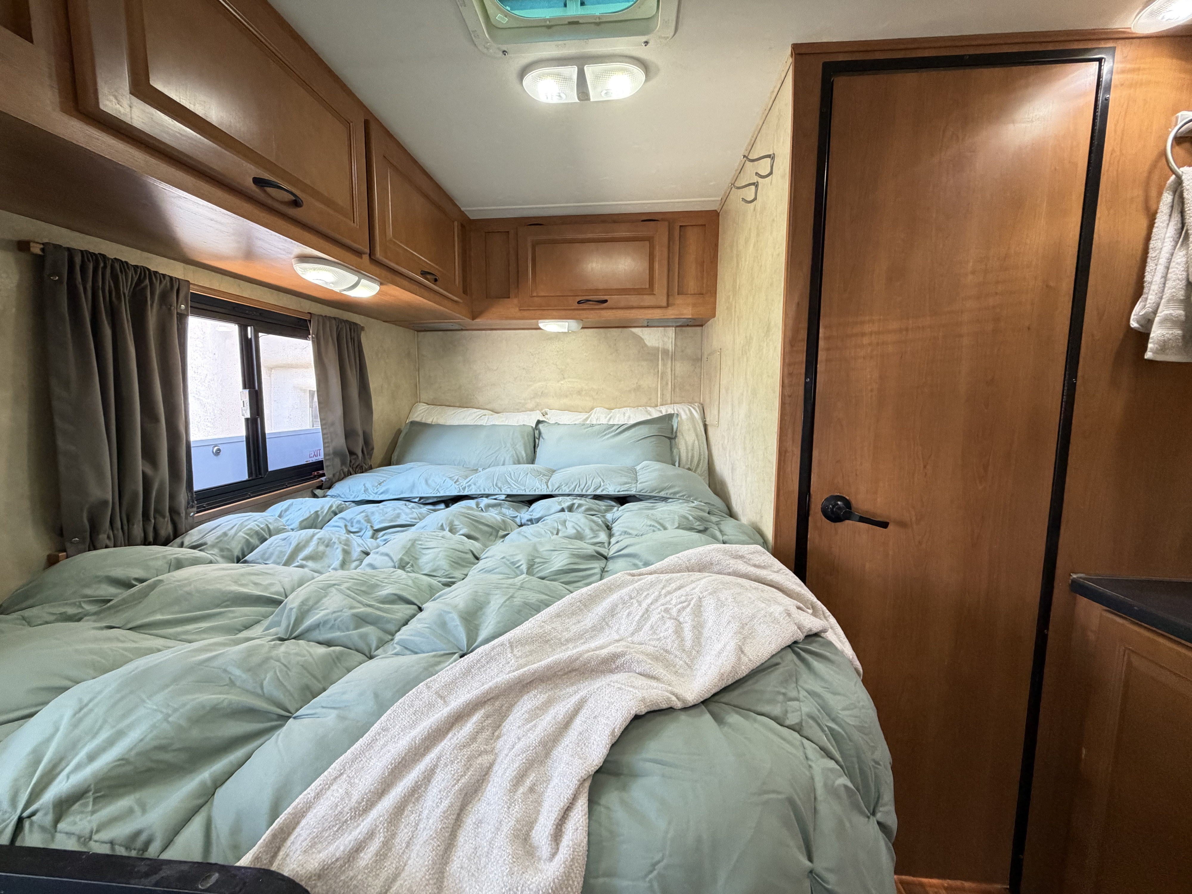 Cozy RV rental bedroom with light blue bedding, wooden cabinets, and a window with dark curtains.. Thor Majestic 2013