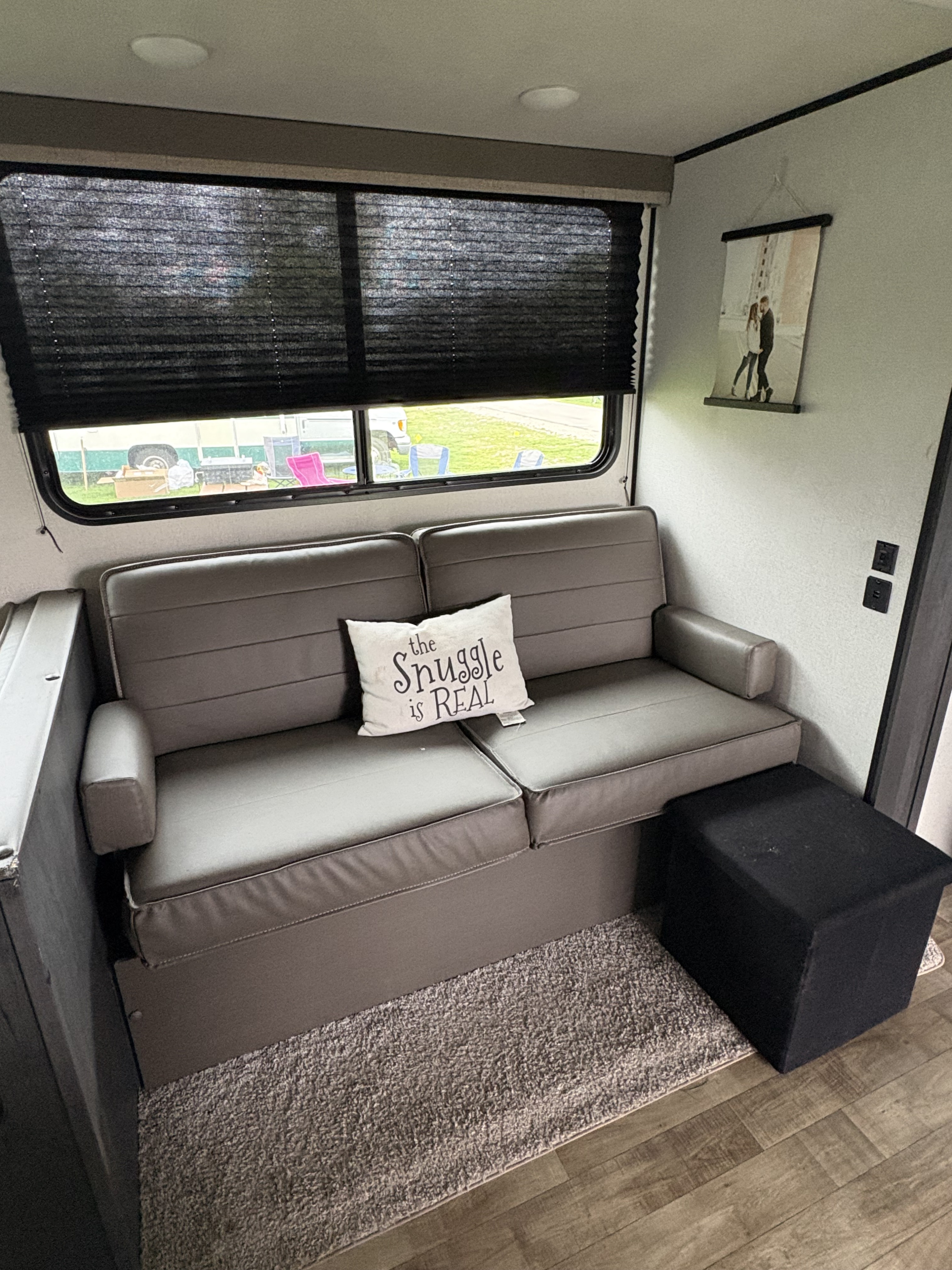 Cozy RV rental living area: gray sofa with 'Snuggle is REAL' pillow, window views, and a black ottoman on a rug.. Keystone RV Hideout 2022
