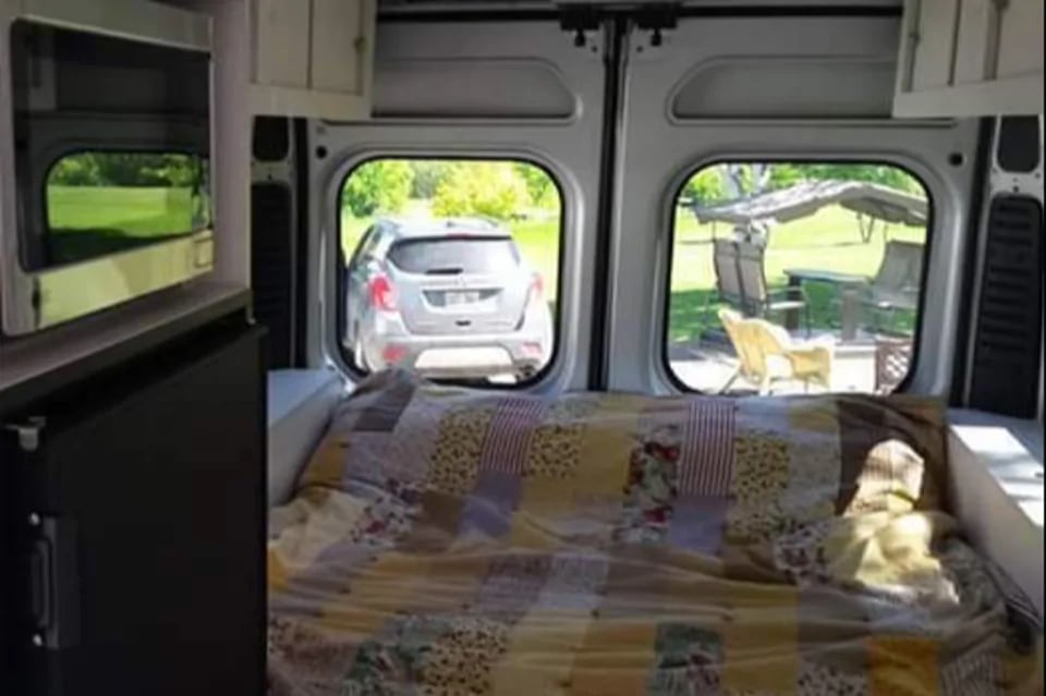 Interior of an rv rental: a bed, microwave & fridge visible. Rear doors frame outdoor views of a parked car and a patio setup.. RAM Dodge Promaster 2015