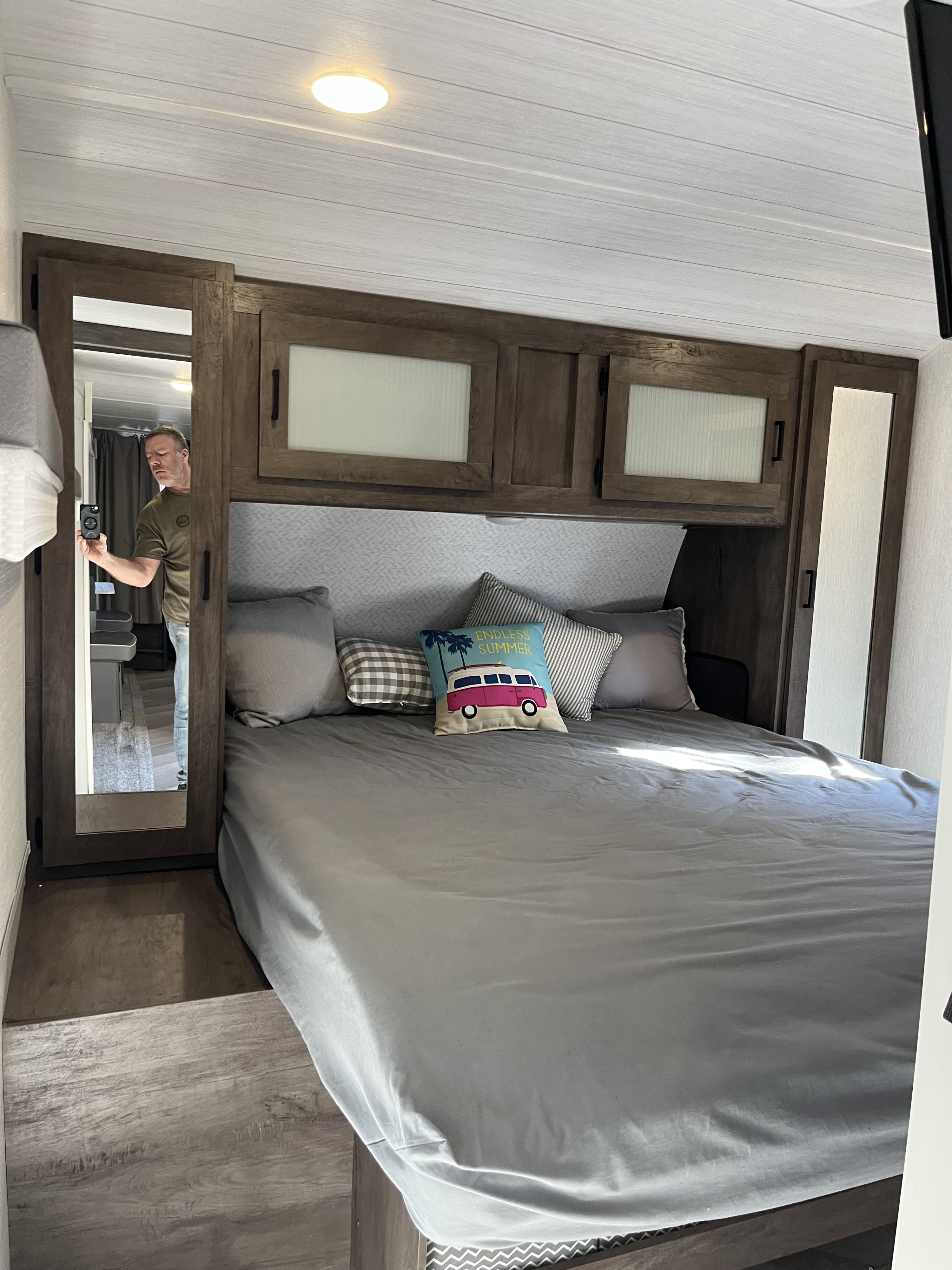 RV rental bedroom with a cozy bed, overhead cabinets, and wood-look flooring. A man is reflected in the mirror.. Forest River Wildwood 2022