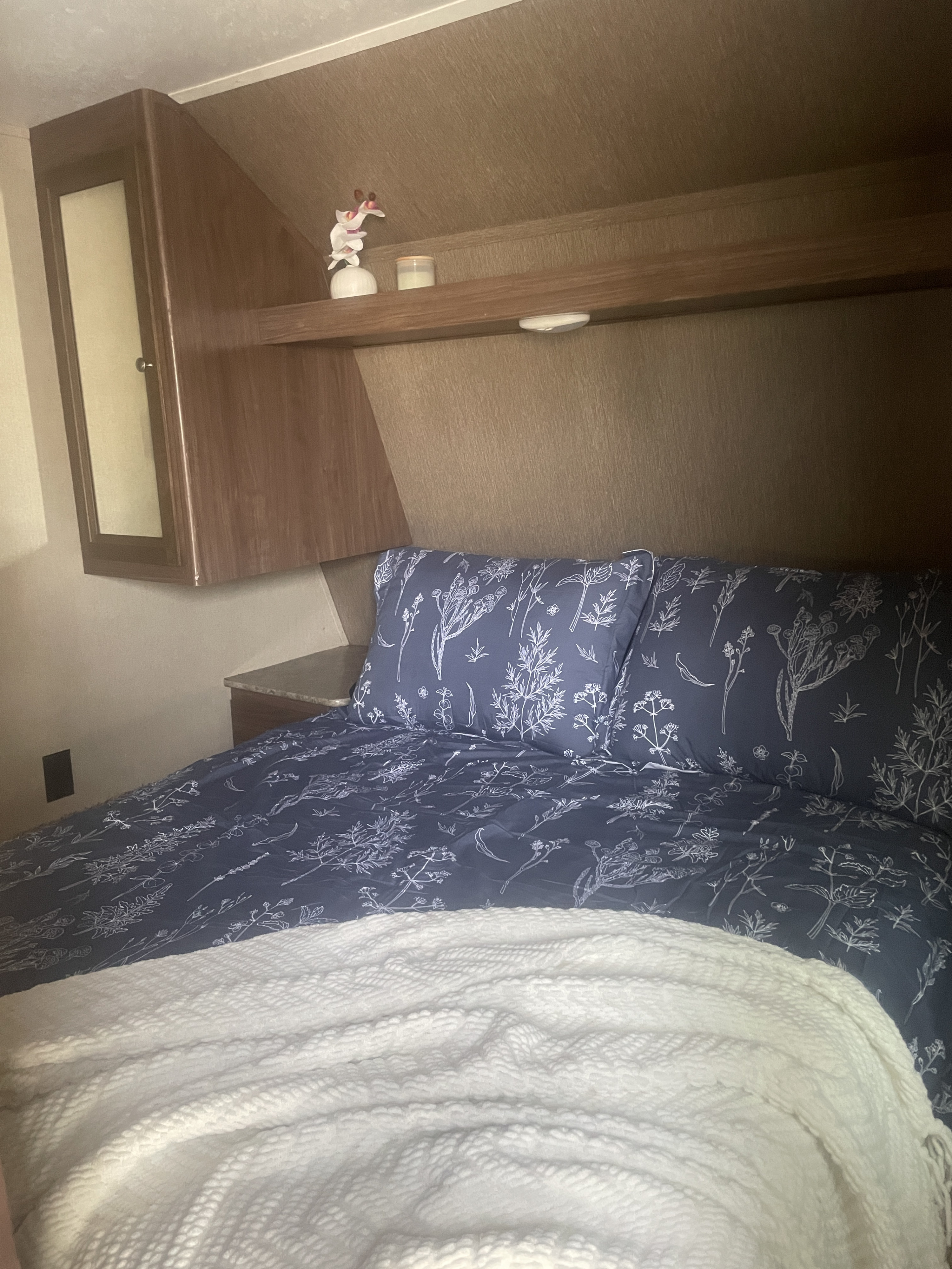 Cozy RV rental bedroom featuring blue botanical bedding, overhead storage, and a fluffy white blanket draped on the bed.. Dutchmen Coleman Light 2016