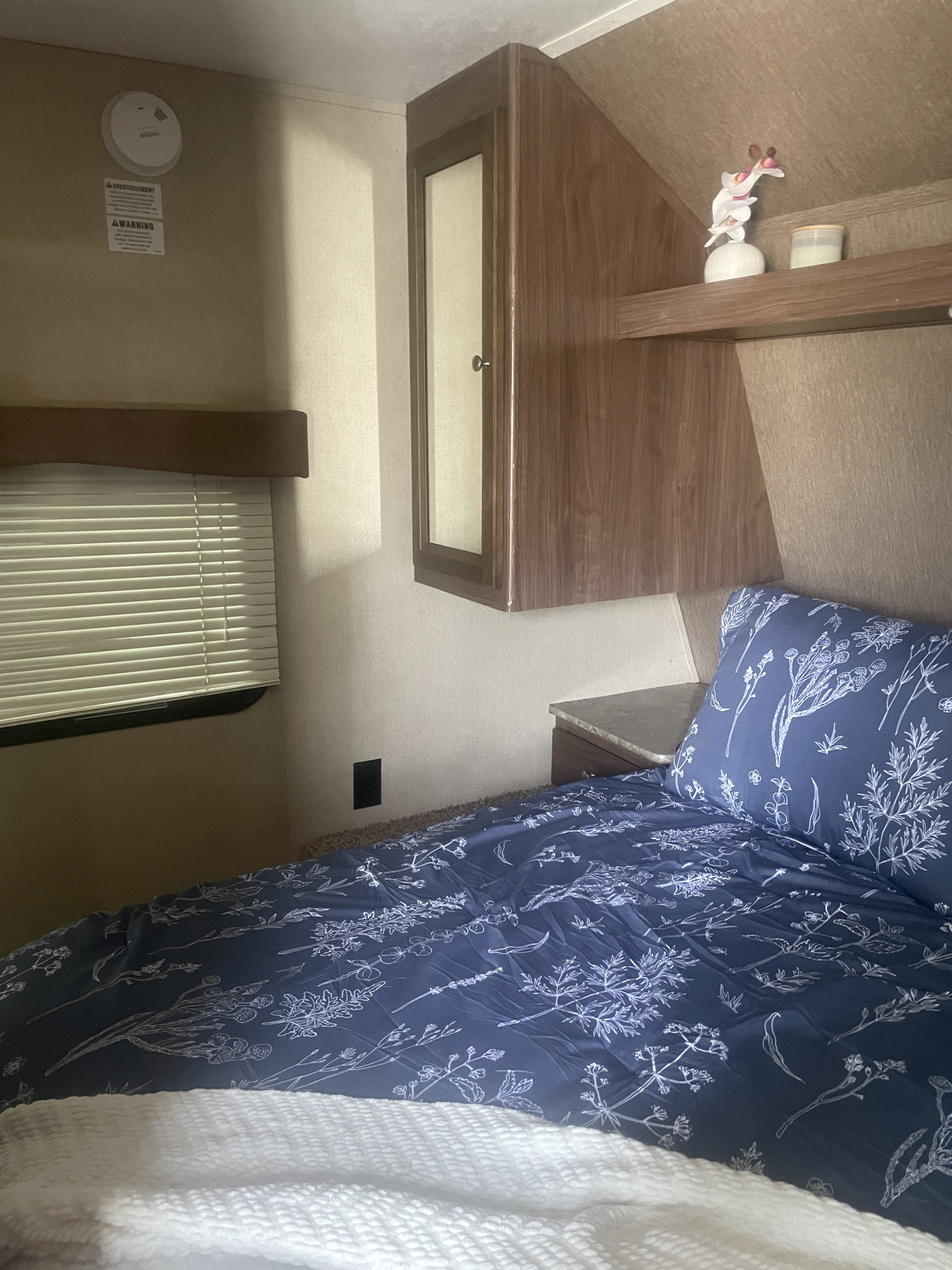 Relax in this RV rental's cozy bedroom. Features include floral bedding, overhead cabinets, and a window with blinds.. Dutchmen Coleman Light 2016