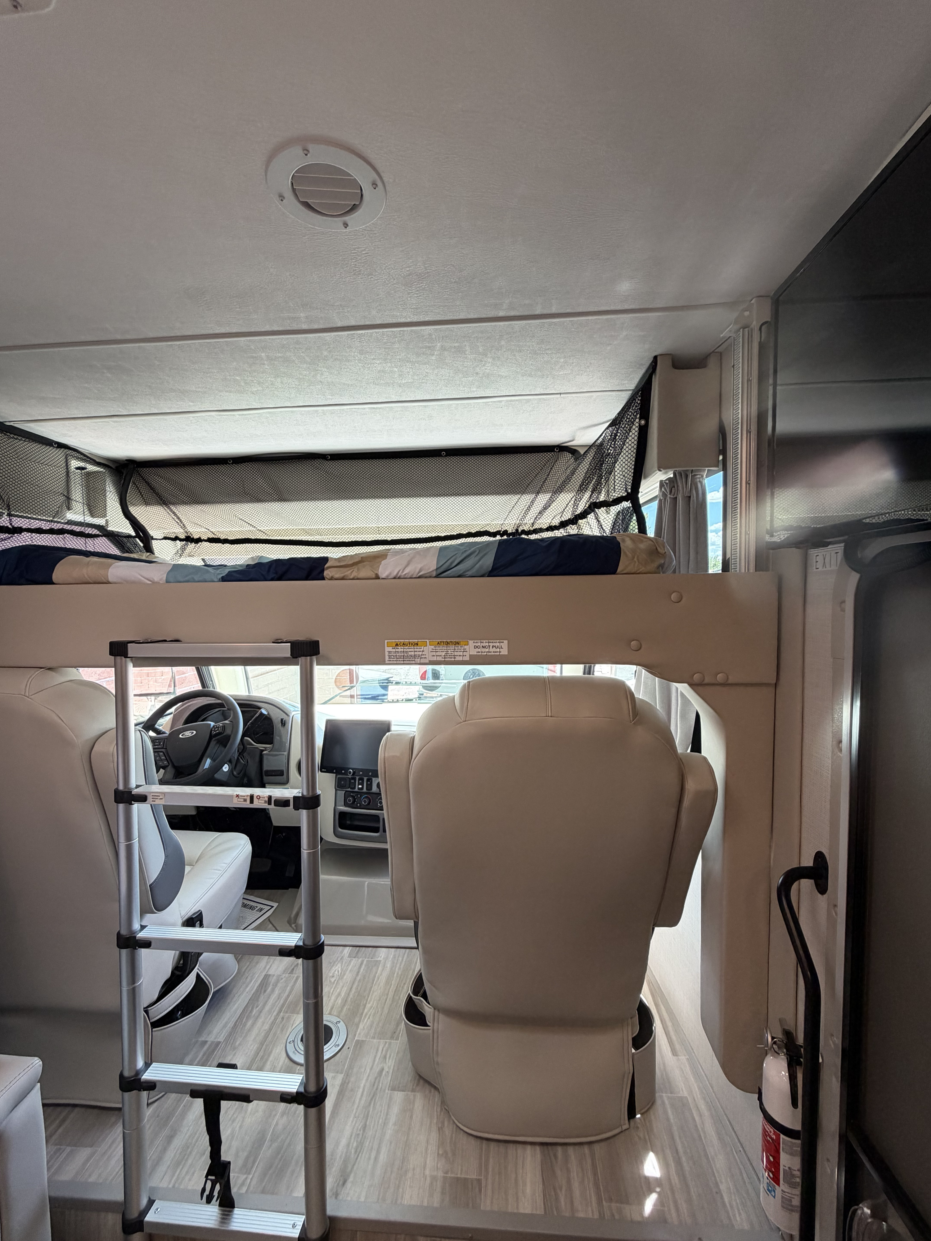 Inside an RV rental: an overhead bunk bed with a safety net and ladder, overlooking the driver's cockpit and passenger seat.. Thor Vegas 2026