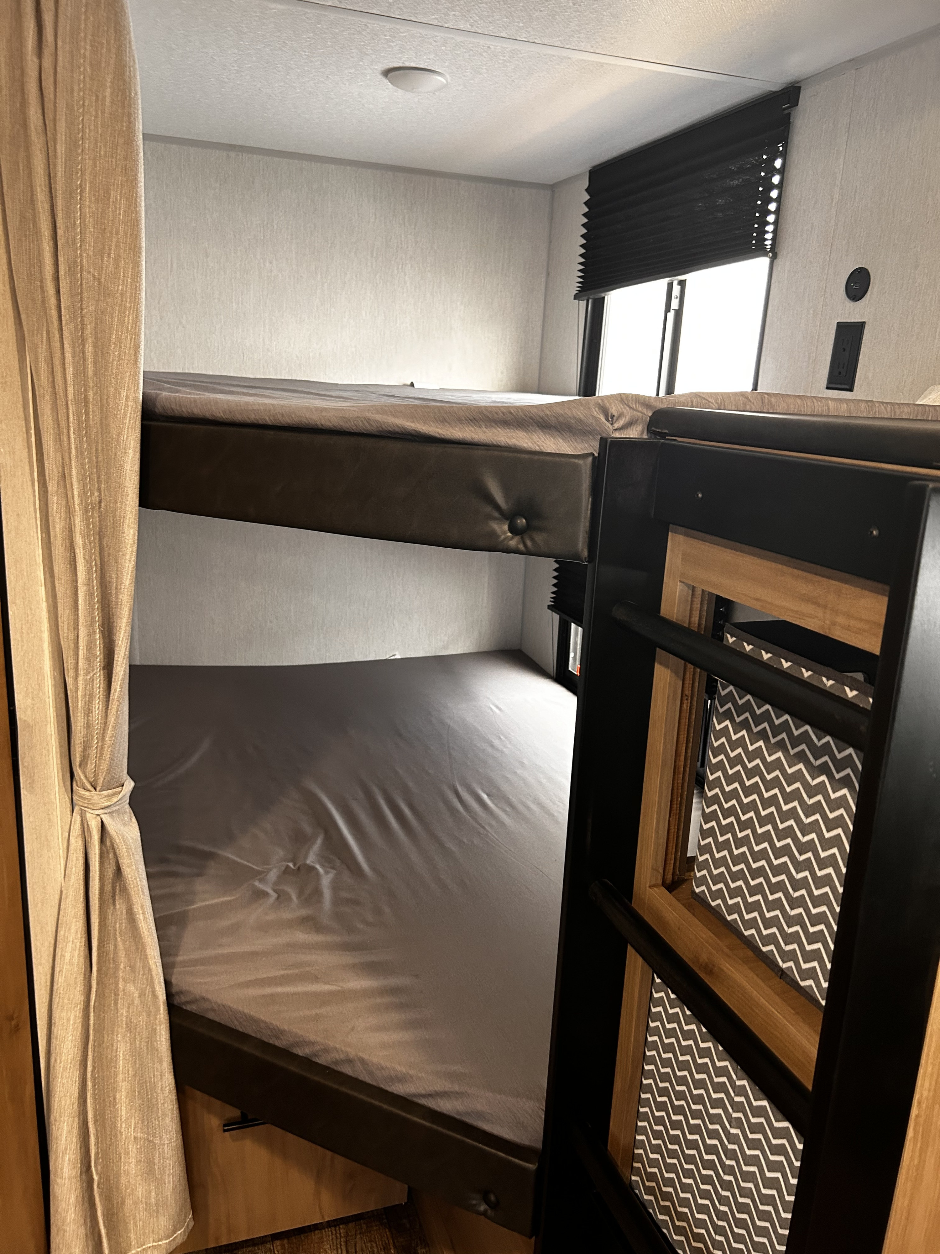RV rental bunk bed setup: Two beds with gray sheets, privacy curtain, window with blind, and wall charging outlets.. Forest River Campsite Reserve 2024
